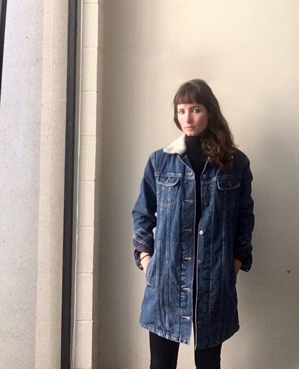 Lindsay stands in front of a white wall, looking at the camera, wearing a long denim jacket