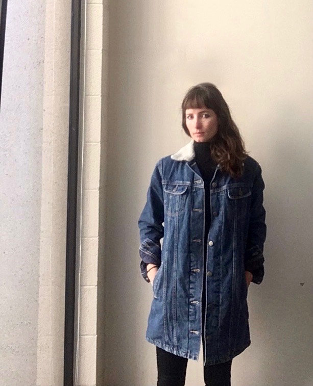 Lindsay stands in front of a white wall, looking at the camera, wearing a long denim jacket
