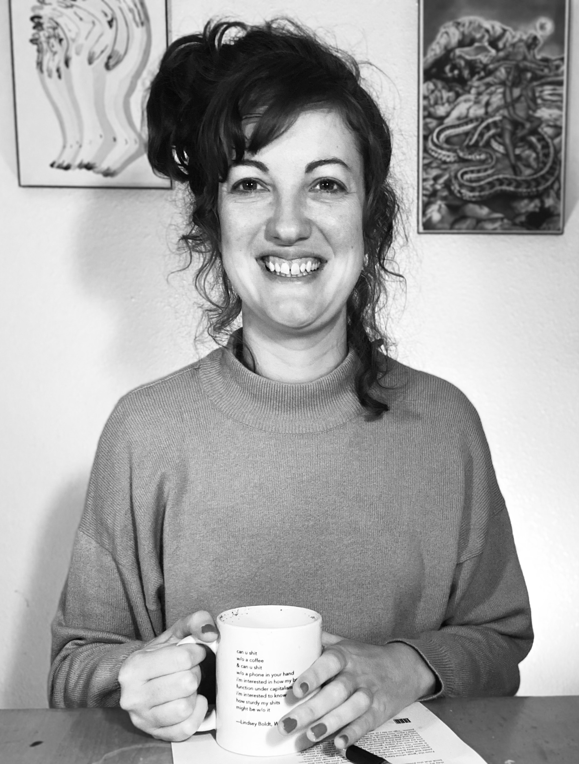 In this black and white photo, Lindsey Boldt sits at her kitchen table, wearing a turtleneck sweater and holding a white mug with a poem from her book Weirding printed on it. The type is too small to read, but it says: "can u shit / w/o a coffee / & can u shit /w/o a phone in your hand / i'm interested in how my bowels might function under capitalism / i'm interested to know / how sturdy my shits/ might be w/o it". She is smiling.