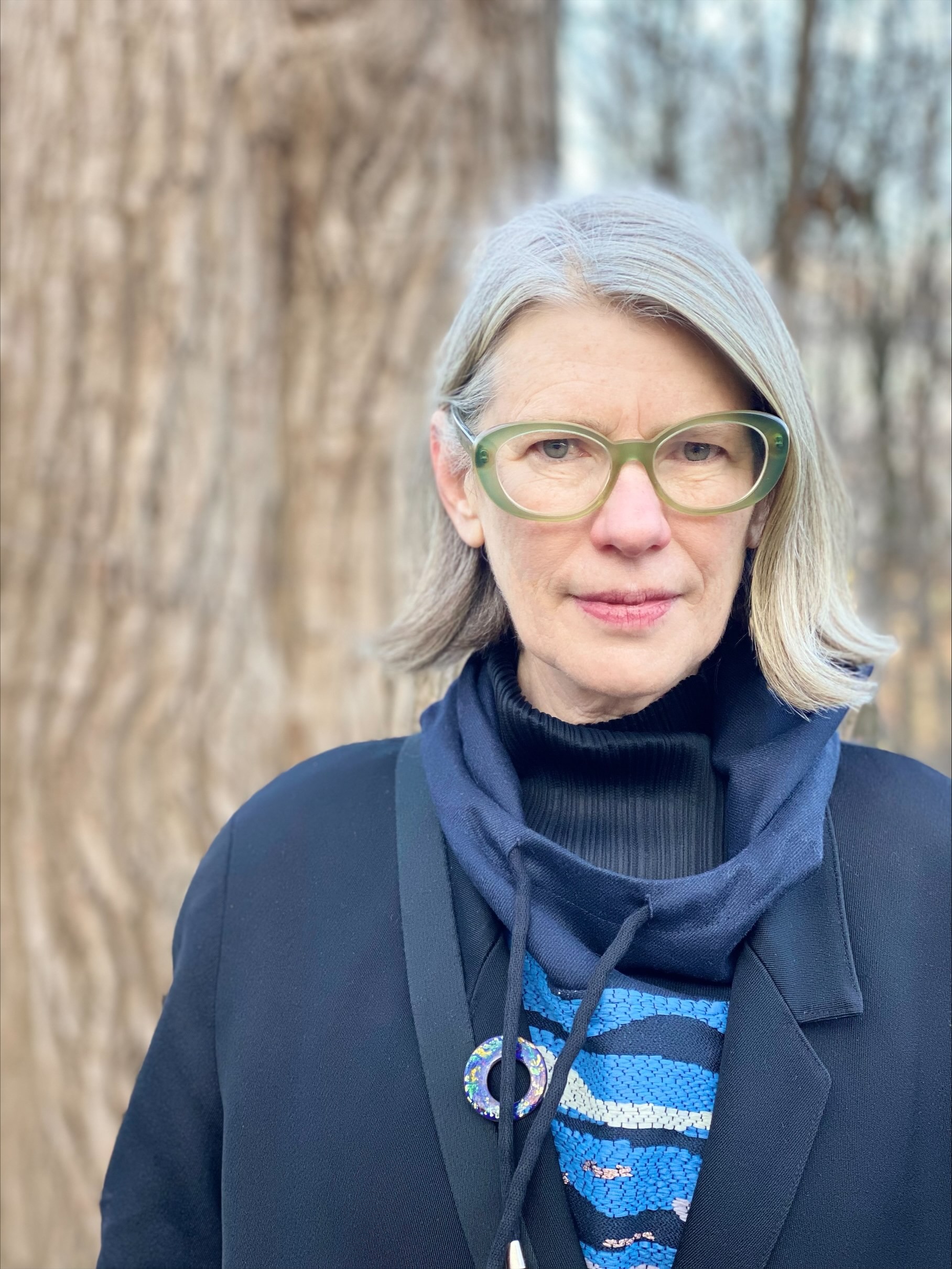 Lisa Robertson stands outside, trees visible in the background but out of focus. She is looking at the camera, wearing green glasses, a dark blue coat, and dark blue turtleneck.