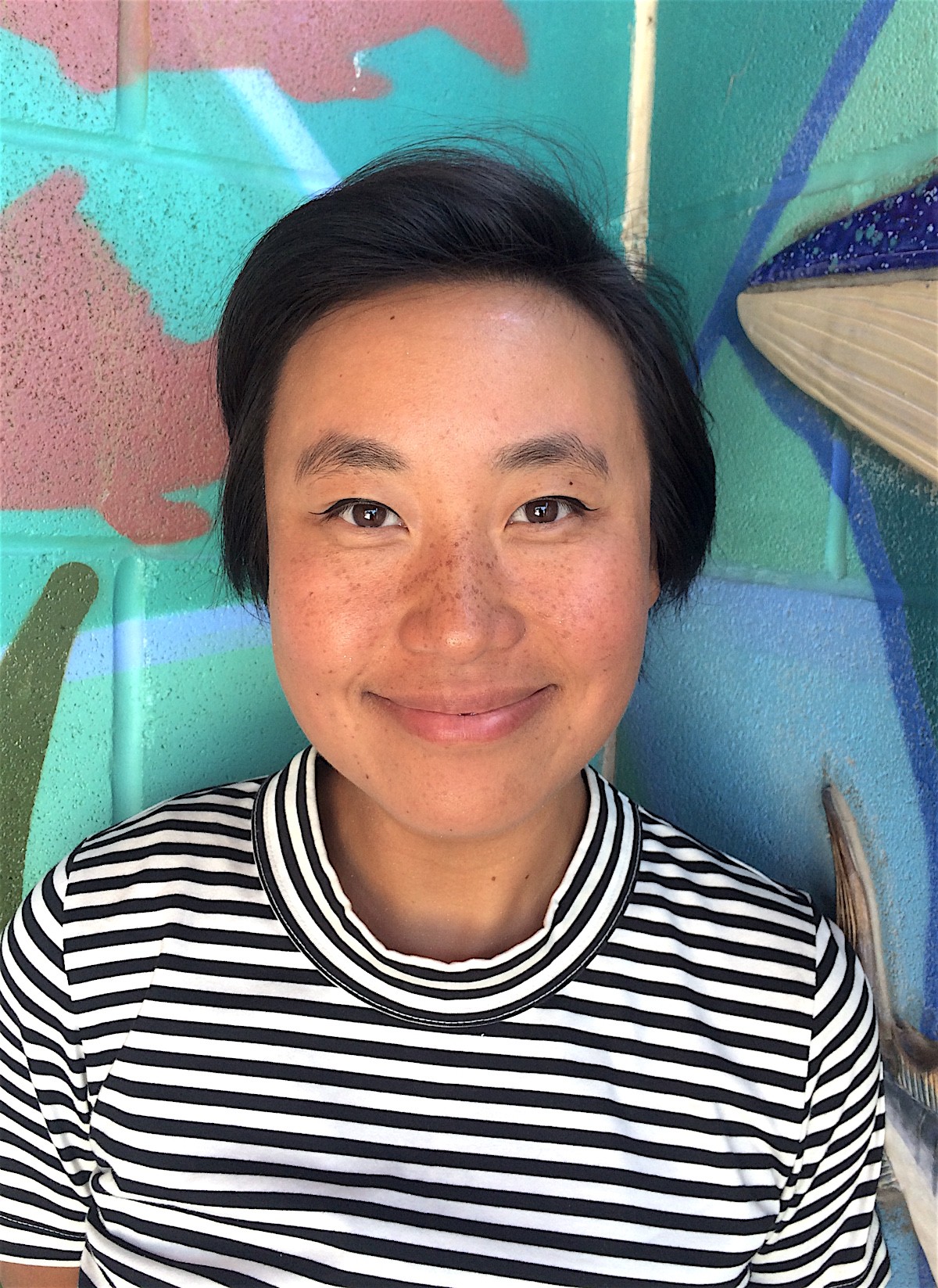 Lynn is centered in the cropped photograph wearing a blue and white horizontal striped shirt with a mock collar. She is smiling and in front of a brightly-painted brick wall.