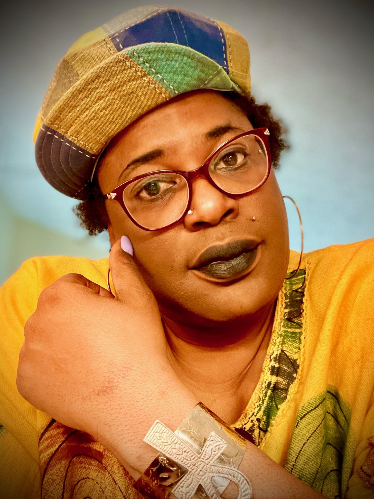 MoHagani squarely faces the camera in a tightly-cropped portrait-oriented photograph. The image is cropped from roughly mid-shoulder in and from mid-chest up. MoHagani is wearing a bright yellow shirt with an abstracted design that's mostly covered by her right arm with is raised with her hand in a fist up to her shoulder; the abstracted design on the shirt looks like foliage. There is a large ankh bracelet on her raised arm. She wears large enamel glasses, large hoop earrings, dark matte lipstick, and a newsboy cap in quilted squares of brown, yellow, green, and blue.