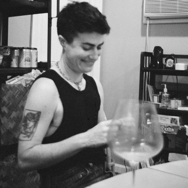 Mack Gregg is featured in a closely-cropped square black-and-white photograph, seated at a table. They are visible from the waist up, wearing jeans and a black tank top and smiling. In the foreground is an out-of-focus wine glass; in the background are black shelves with beer, hand sanitizer, and various objects on them.