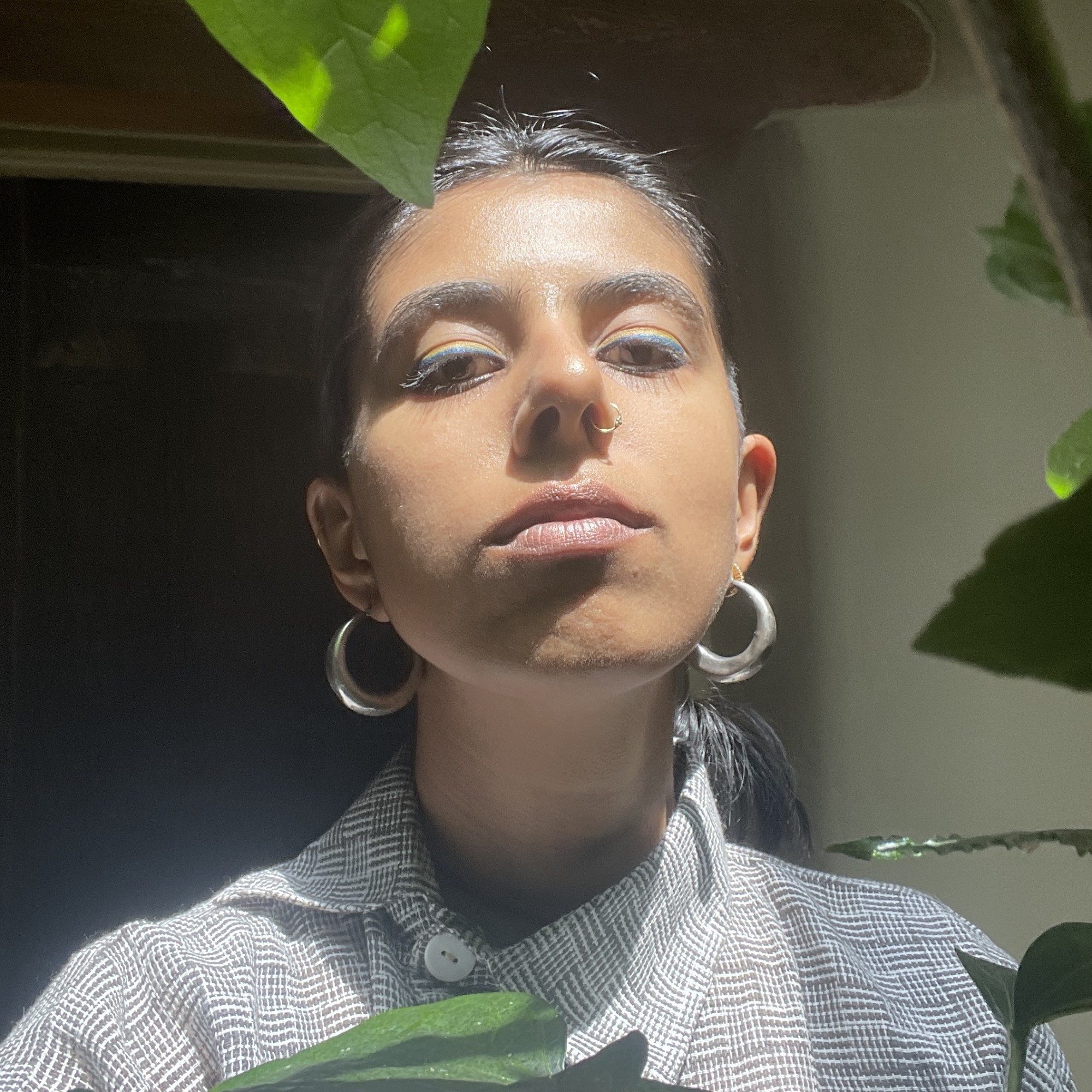 Mallika Singh is standing in bright light next to a plant with large green leaves, wearing hoop earrings.