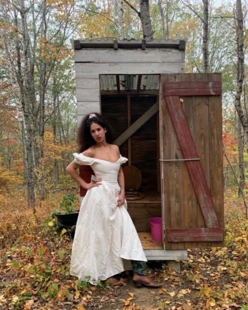 Manal Kara is wearing a floor-length white gown, standing in front of a wooden outhouse on a fall day.