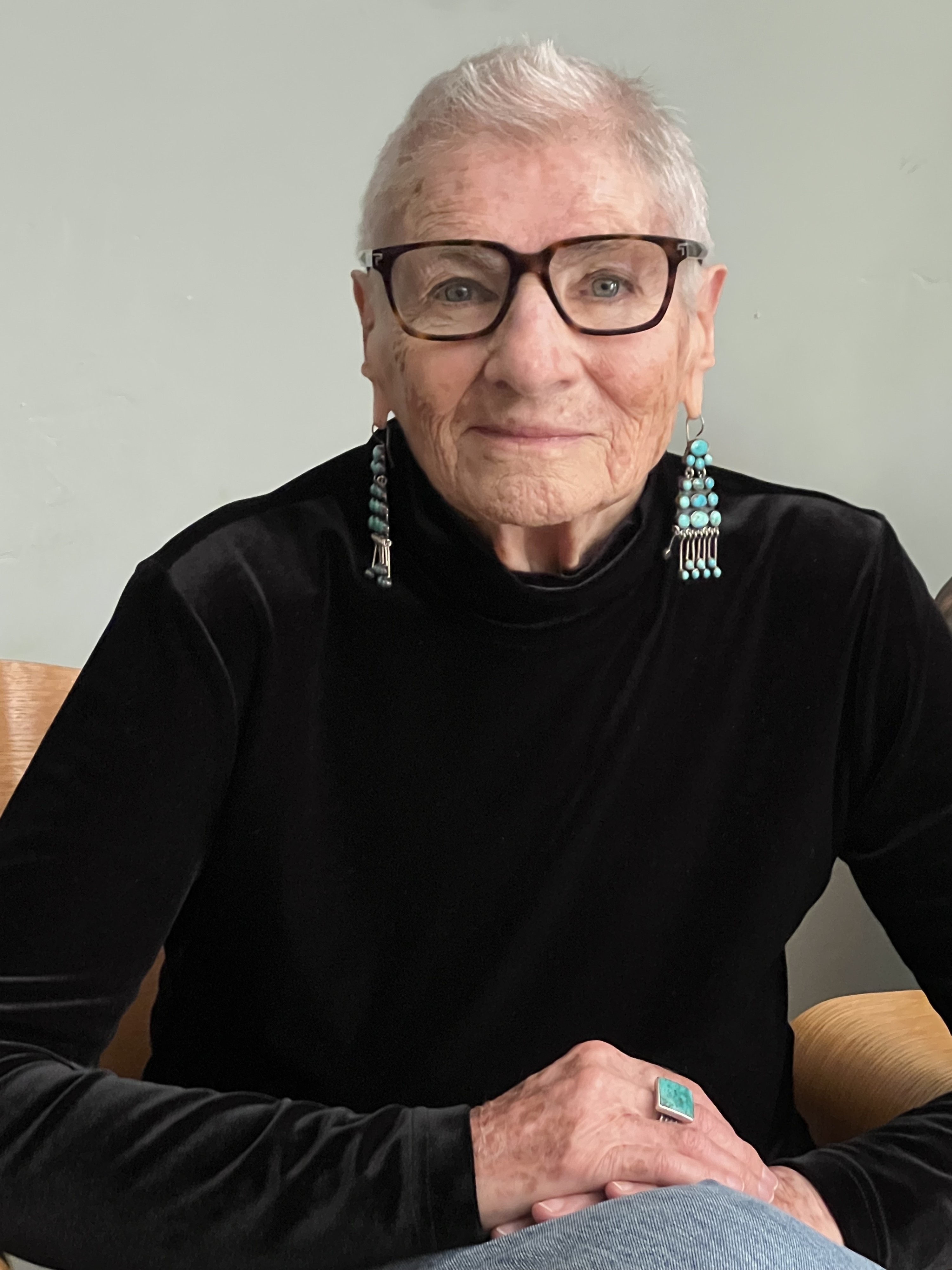 Margaret Randall is shown in a color photograph cropped tightly from the waist up. A small patch of green in the background suggests that she is seated in an armchair. She is wearing a black velvet long sleeve shirt, large dangly earrings, and is looking directly into the camera and smiling subtly.