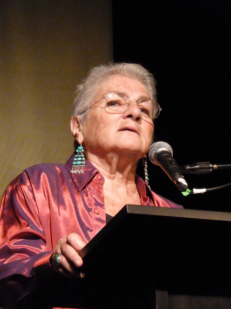 Margaret Randall is photographed speaking into a microphone at a podium wearing turquoise earrings.