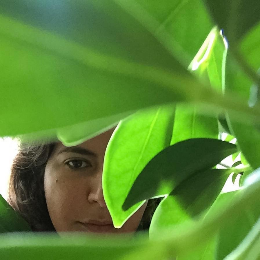 Marwa Helal stands behind a plant with large green leaves, her face partially obscured the its leaves.