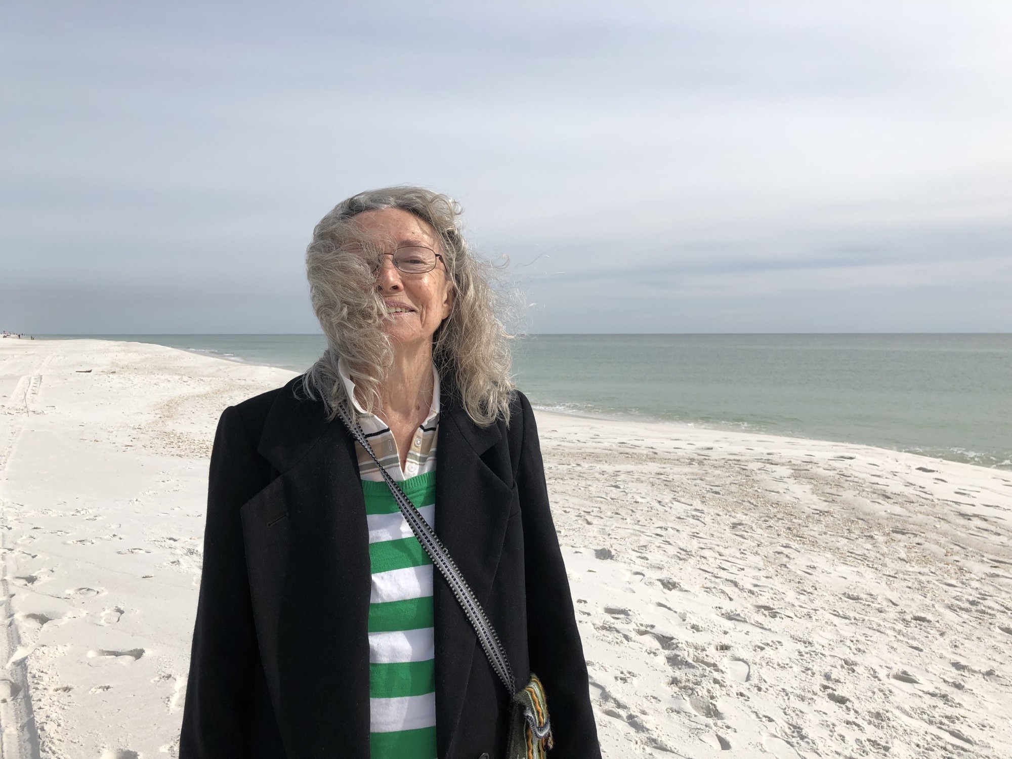 Poet is standing on a wide, white sand beach with blue/green ocean close-beside to her left and as horizon behind her.  Sea breeze blowing poet's hair forward and partially covering her face.  Poet is wearing a black winter coat over a light sweater with wide horizontal green stripes. A small, colorful cloth and woven Peruvian bag is slung over her right shoulder and across her torso.  Poet is slightly squinting from hair and sun and smiling.  A sky layered of blues and paler washes of blues and way-subtle pinks spread and fill the space above the water.