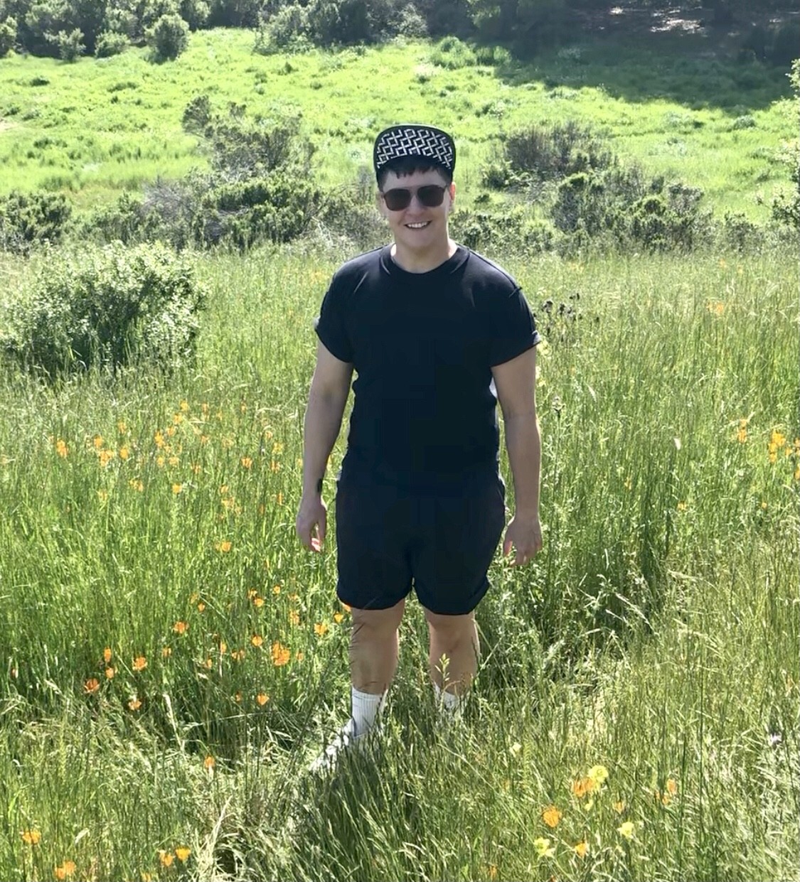 Maxe Crandall stands in a field of wildflowers, looking at the camera and smiling, wearing all black with a black and white cap.