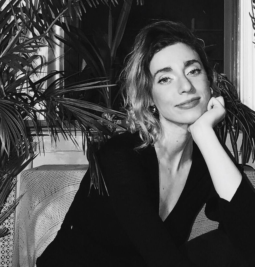 the photo, rendered in black and white, shows Maya cropped from the waist up, seated in the right of the frame, resting her chin on her left hand. To the left of the frame is a large house plant.