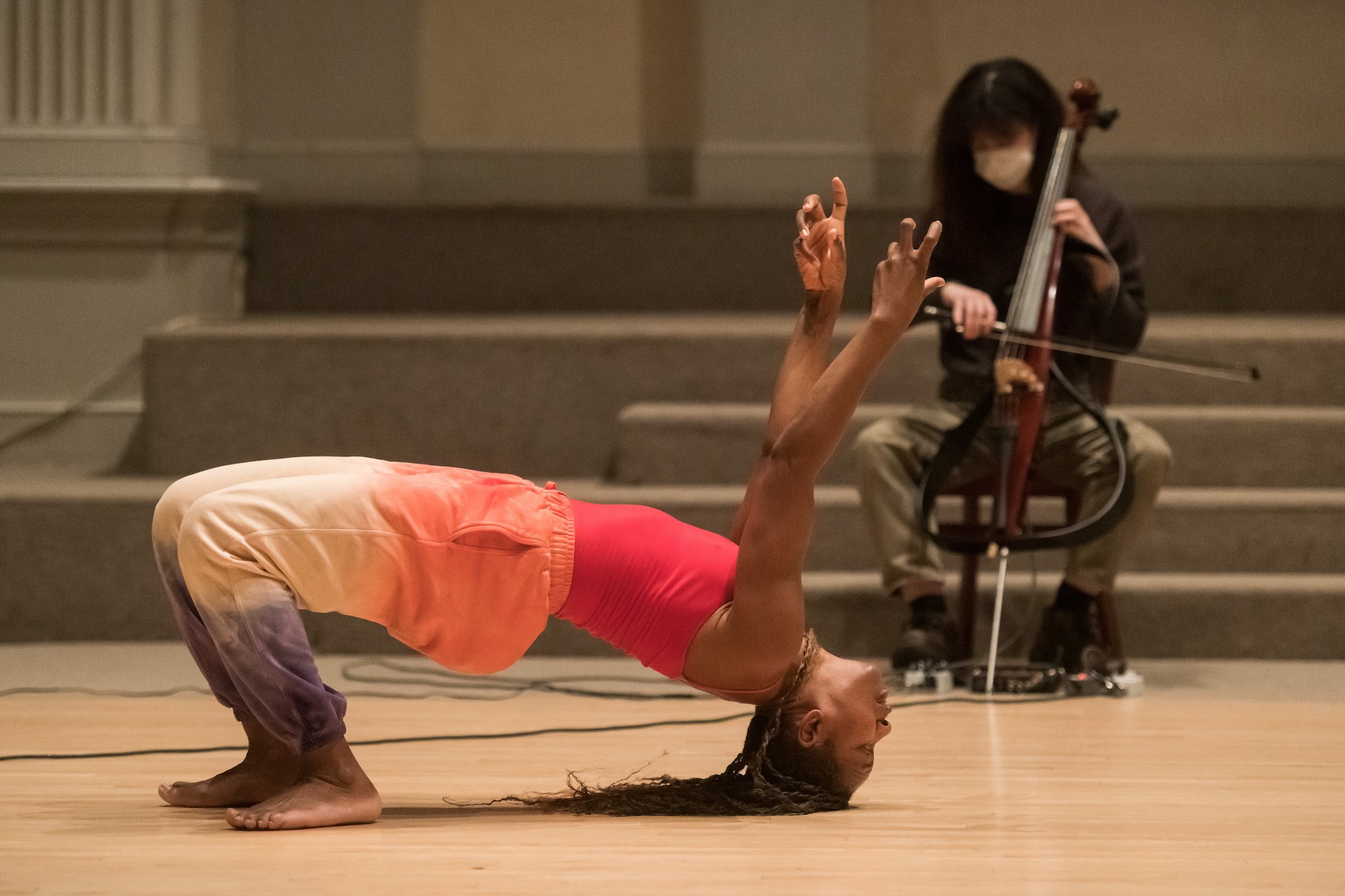 mayfield, a brown-skinned person with long brown braids, is in the foreground of the photo in a church with an altar and electronic cellist in the background. mayfield is in a backbend position with their head and bare feet touching the blonde wooden floor--arms stretched up towards the sky & wearing pink and purple sweats and a red  tank top. mayfield's fingers are curled & expressive. The cellist is blurred, wearing a black sweater, brown pants, and black boots.  The carpet on the stairs of the altar behind the cellist is a tan color. The cellist, named Dorothy, is sitting on a chair in front of the church altar stairs & is a light brown skinned person with long hair and bangs wearing a white mask & playing their electric cello with cables attached to it.