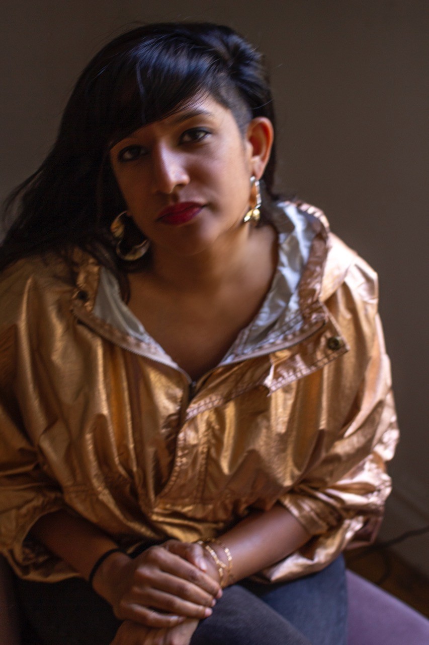 Megan Fernandes wears a shiny gold jacket, large gold hoops, and red lipstick. Her hands are in her lap and she's looking at the camera.