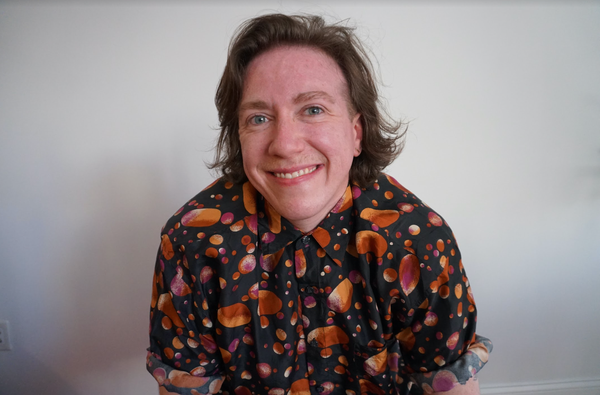 Megan, a white nonbinary person with shoulder-length brown hair, is inside, pictured against a gray wall. They are smiling and wearing a black button-down silk shirt with a celestial potato pattern.