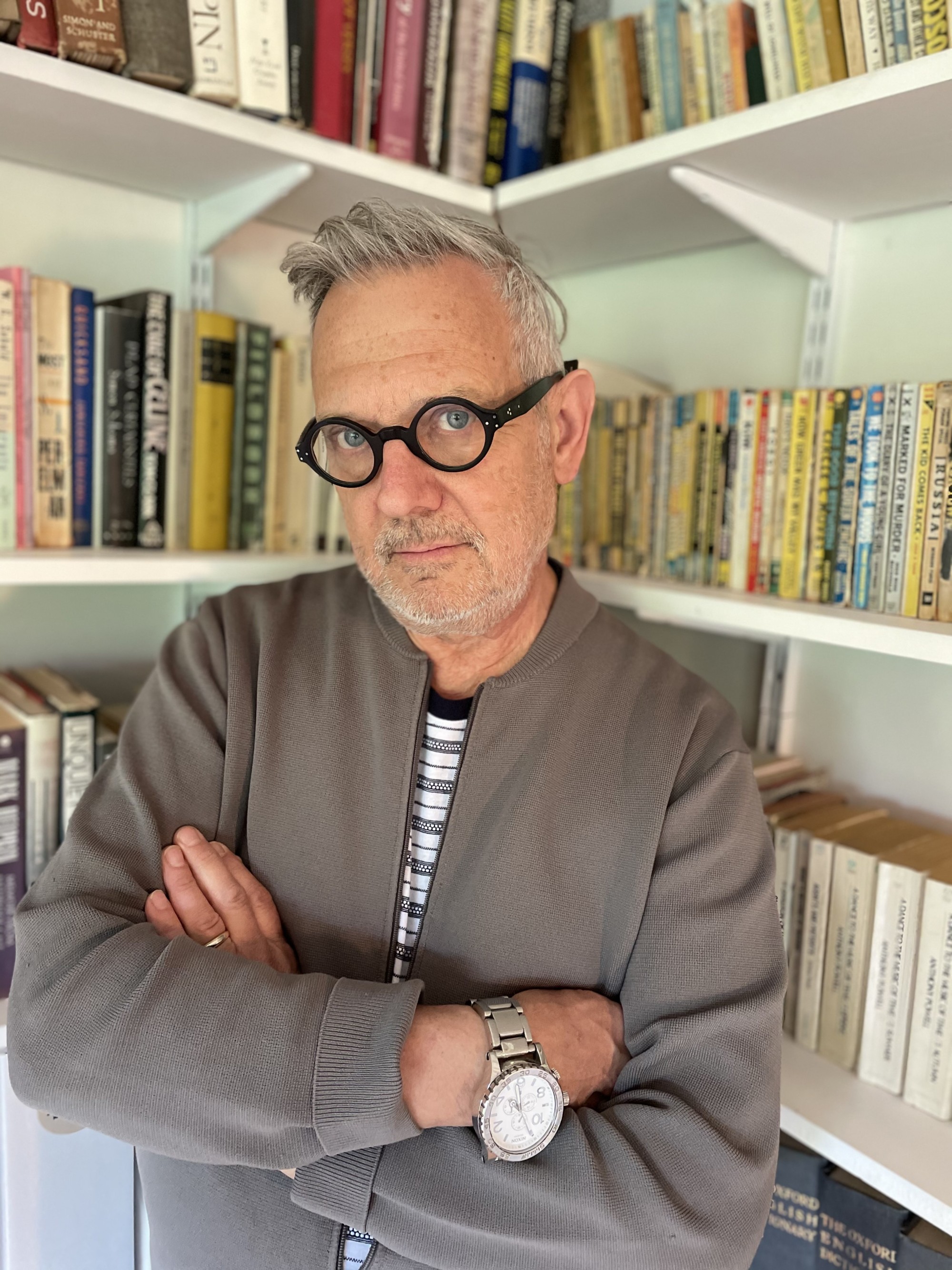 Michael Gottlieb stands with his arms crossed at a corner where two bookshelves meet, wearing a gray jacket and black glasses