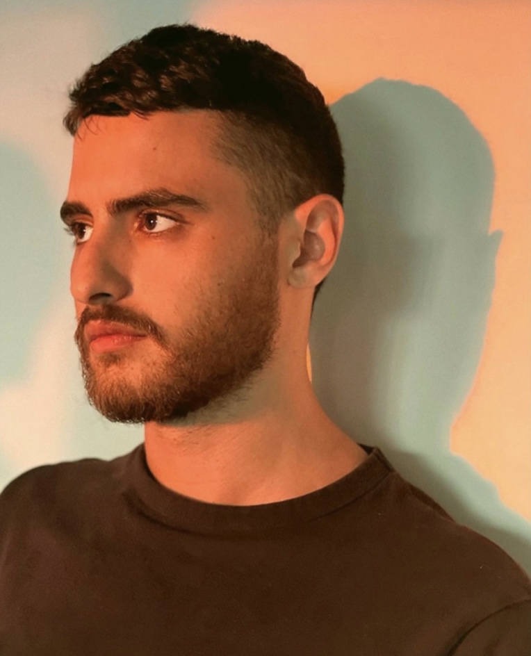 A bearded man in a black shirt stands against a plain background, suffused with orange light.
