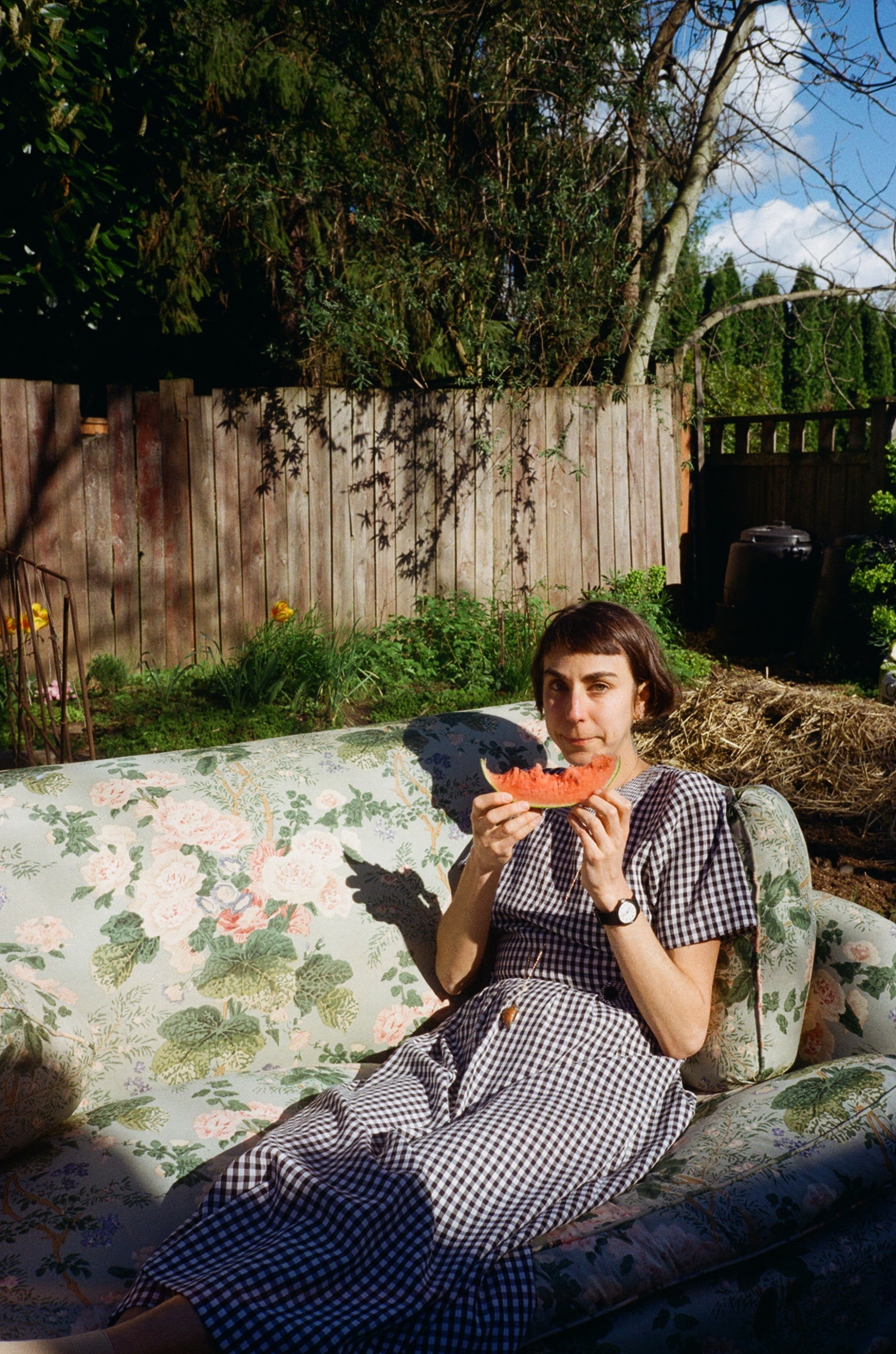 Molly Schaeffer is featured in a portrait-oriented photograph, outside on a floral couch, reclining upright on the couch with her legs coming towards the at an angle. She wears a sundress and is holding a piece of watermelon with her two hands. Behind is a yard with a tall wooden fence, bright green grass and trees, and in the right corner, blue sky and clouds.