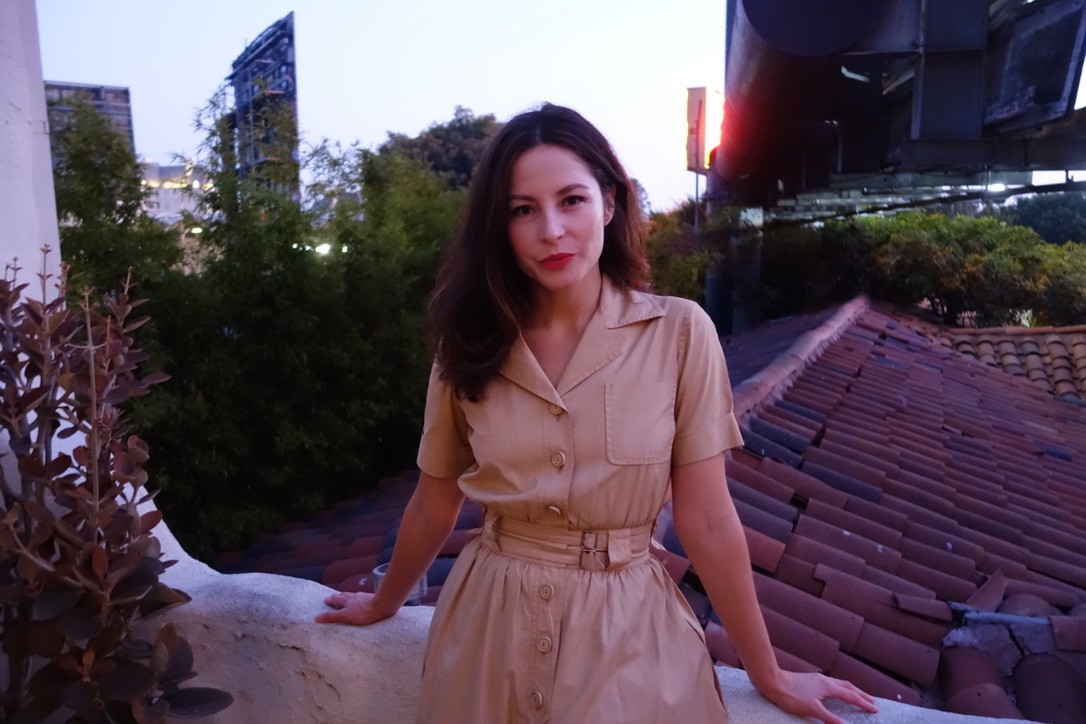 Monica McClure is centered in the picture, wearing a tan trench-style dress, resting on a balcony facing the camera. Behind is a terra cotta roof, trees, and billboards.