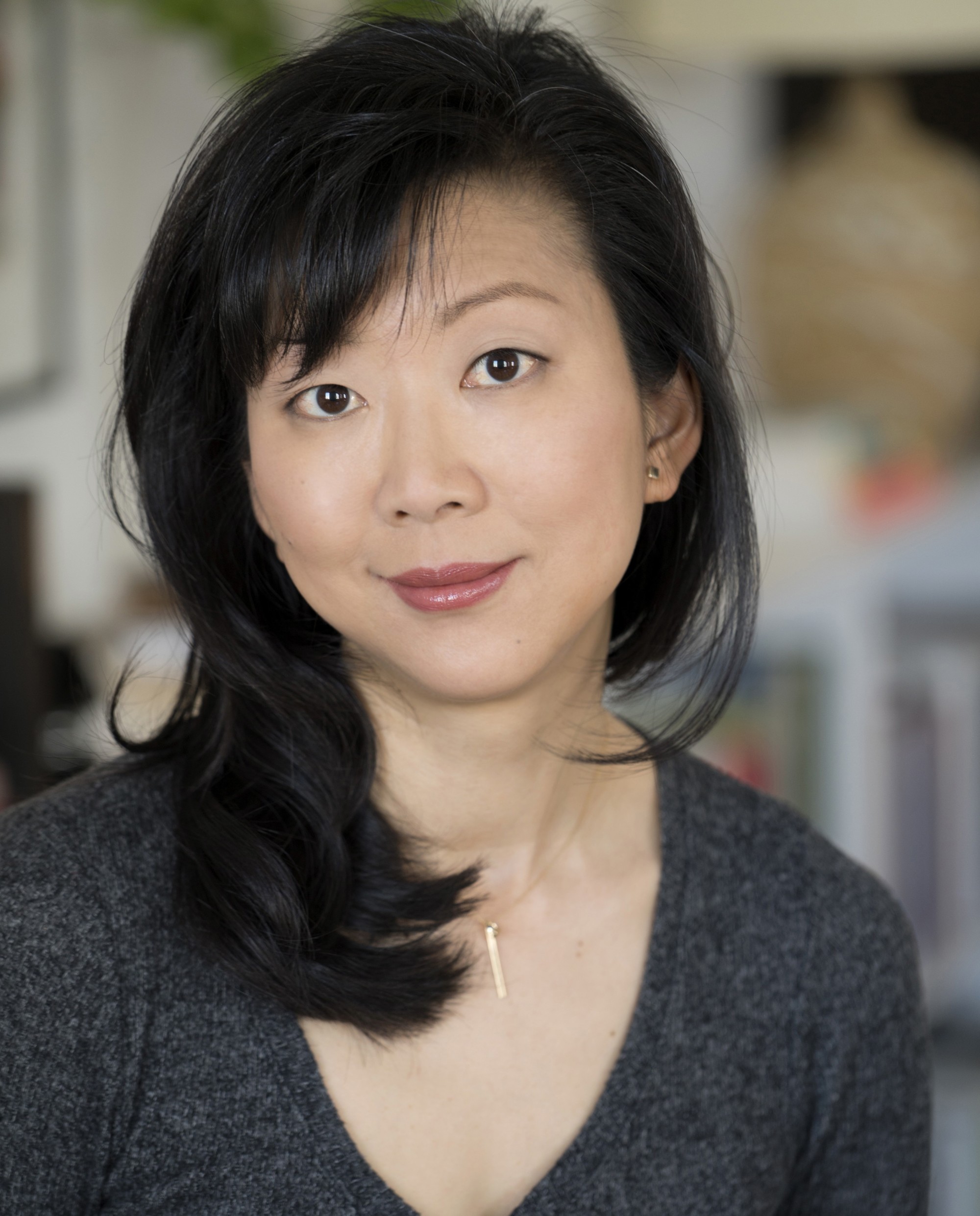 The portrait is cropped from the chest up; Monica Youn is featured center, smiling, wearing a grey heathered shirt and gold necklace. The background of the portrait is out of focus.