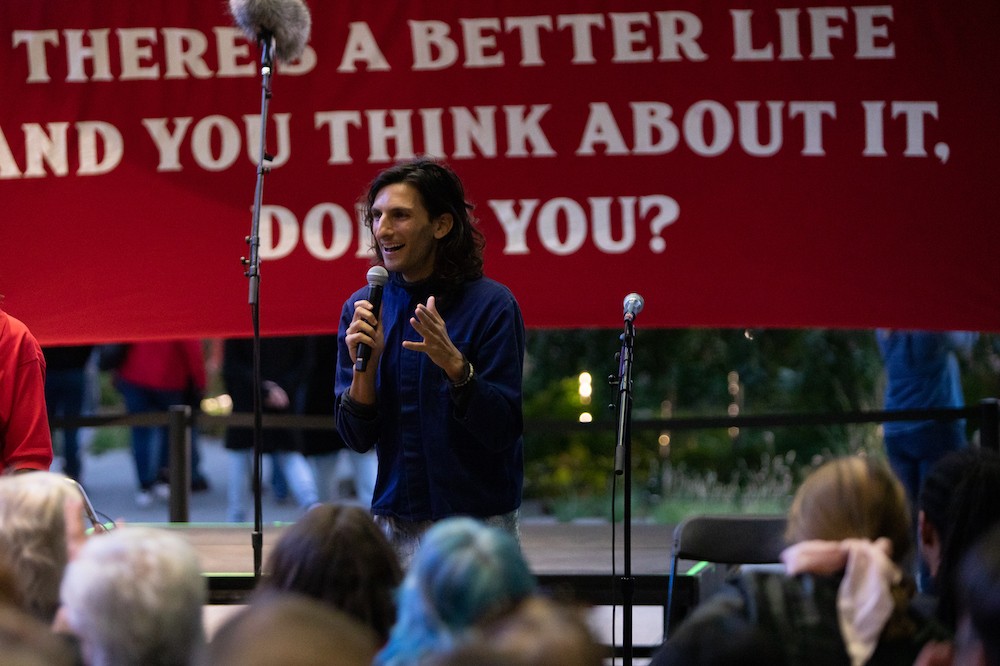 Morgan Bassichis is holding a microphone, speaking to a crowd. They are standing in front of a banner, which is partially blocked from you. The legible part reads, "THERE'S A BETTER LIFE / AND YOU THINK ABOUT IT / DON'T YOU?"