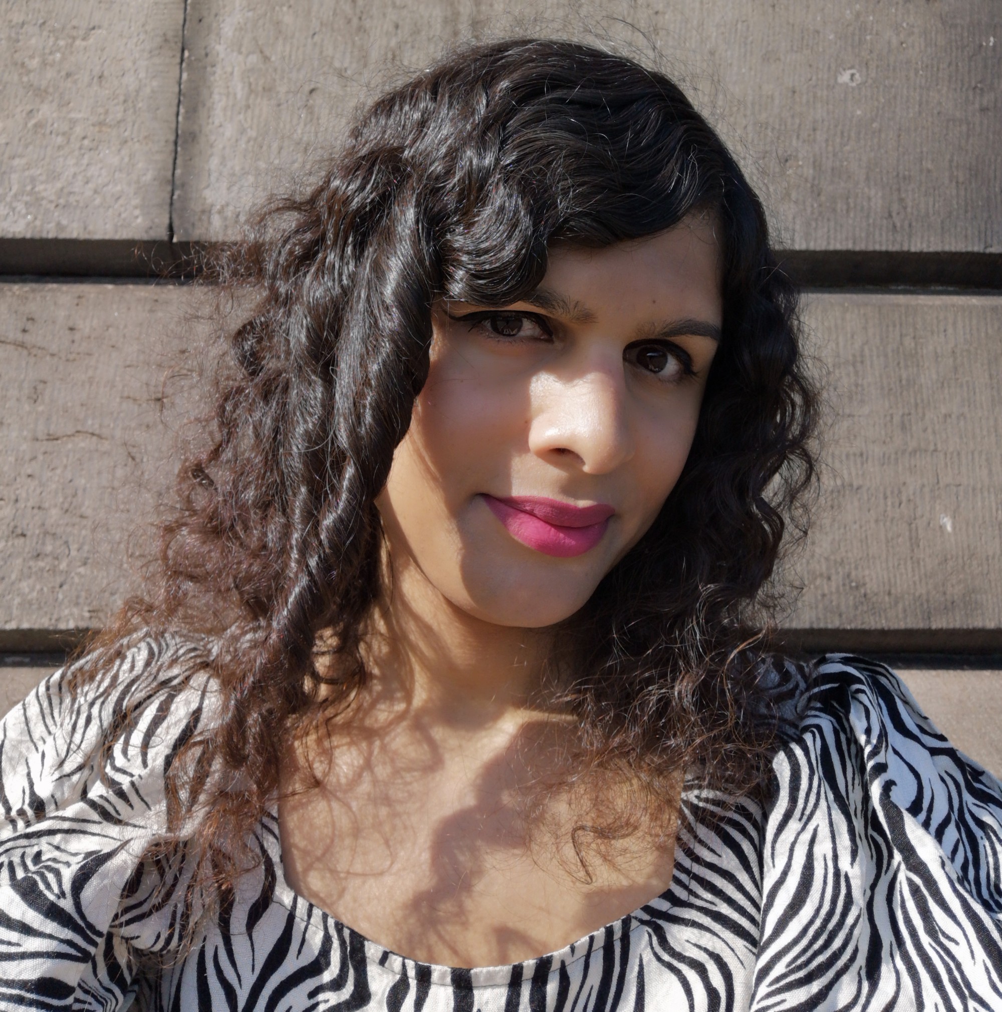 Image of Nat, a brown femme with long curly hair, wearing pinkish lipstick and a zebra-print dress. Her hair casts shadows over her neckline.