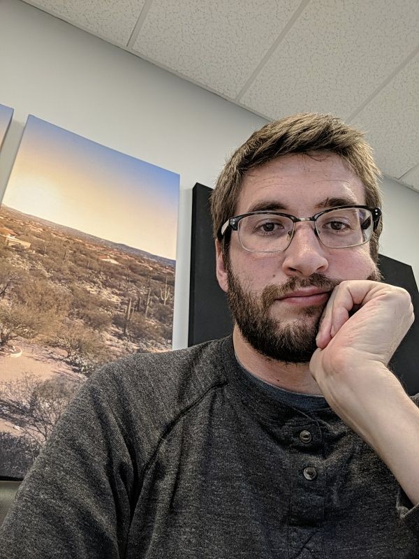 Nate Keen is pictured from the chest up in a green heathered henley, his hand resting on his cheek. In the background is a color photograph of a desert landscape.