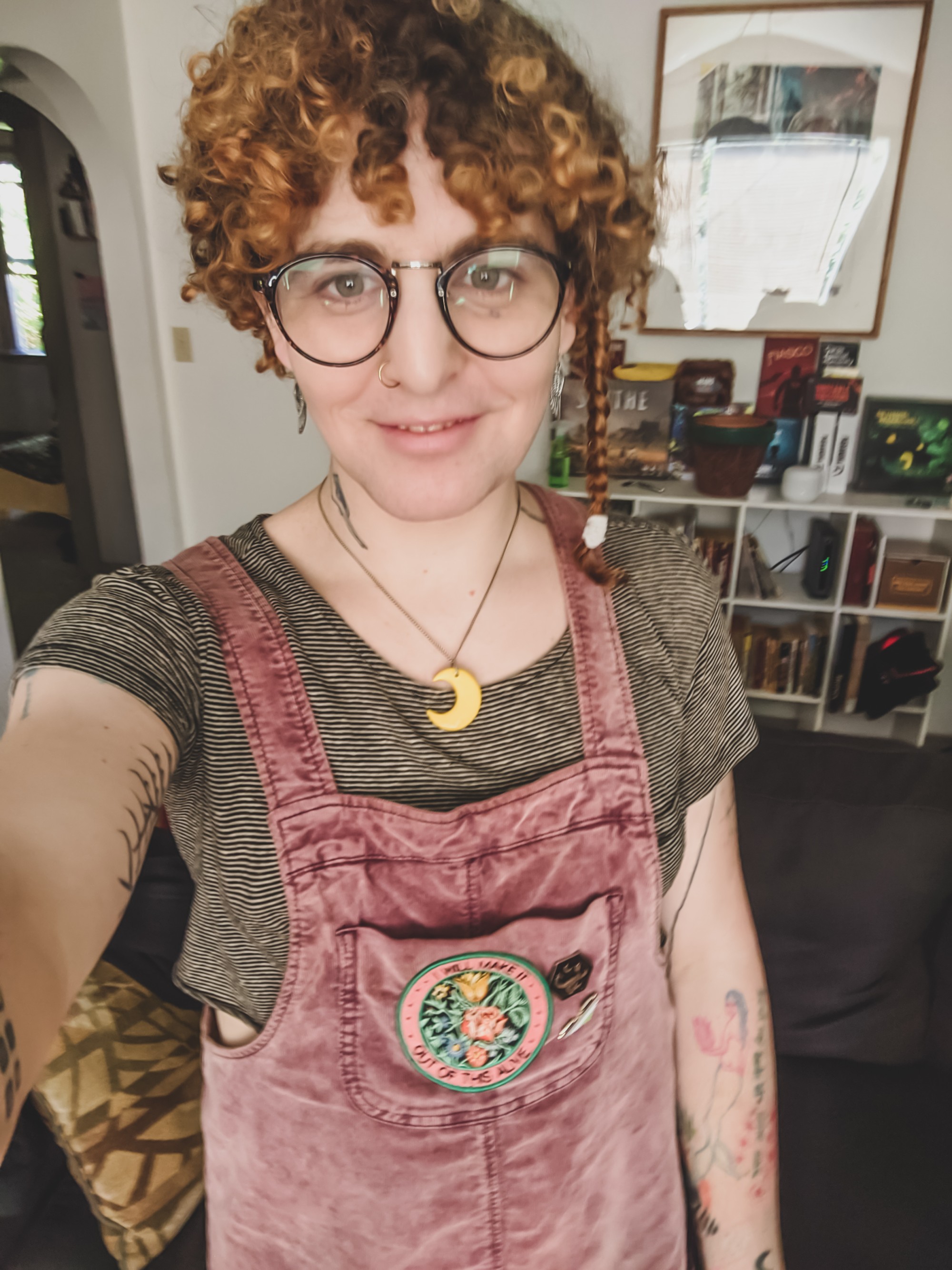 Never Angeline Nørth is shown in a selfie-style photo, holding the camera and centered, cropped from mid-waist up. She is wearing pink overalls with a black and white horizontal striped shirt underneath, a large yellow crescent moon necklace, and large round glasses.