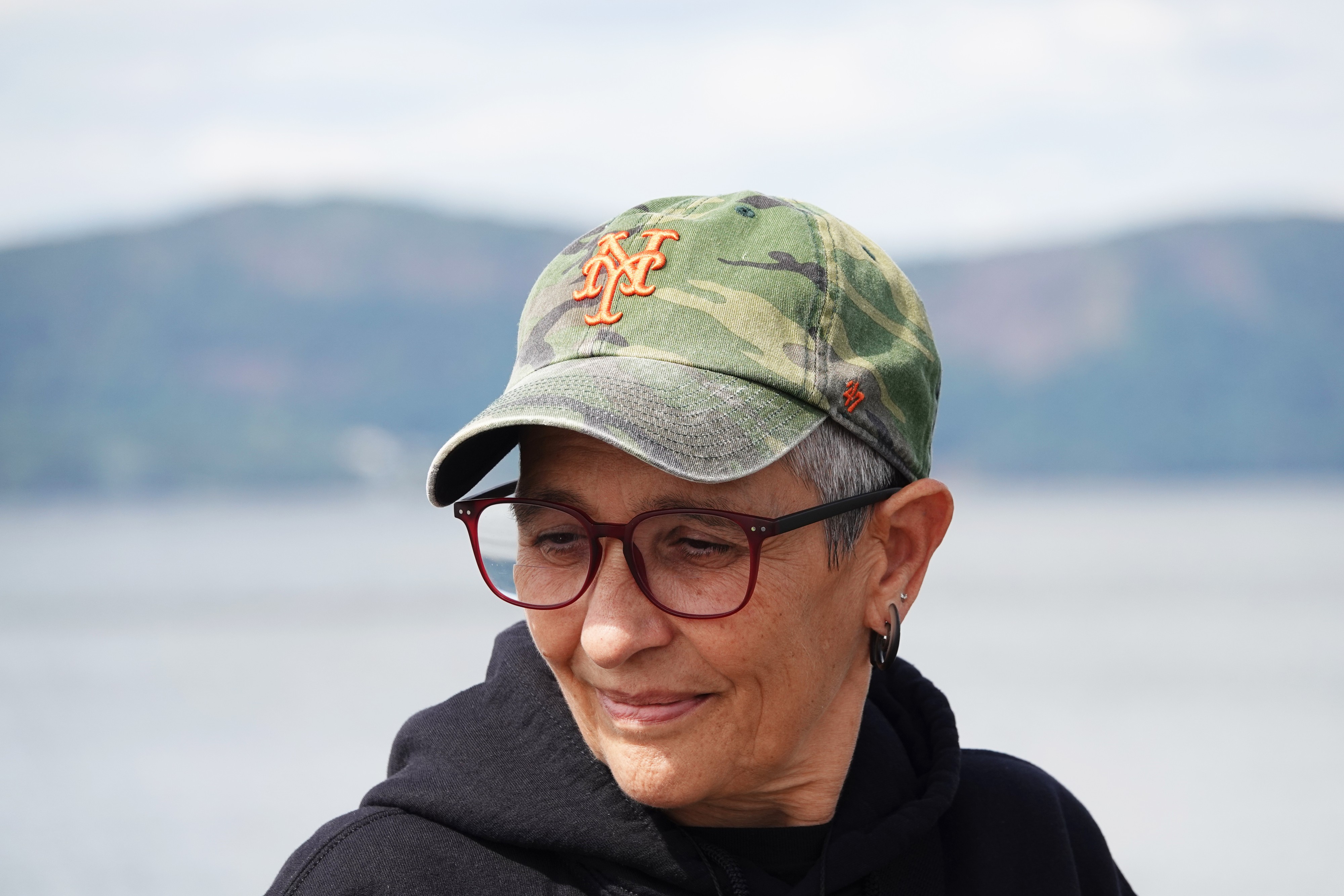 Nicole Peyrafitte is shown in a landscape-oriented color photograph, cropped from the shoulders up. She is wearing a black hoodie, enamel glasses, spiral-shaped earrings that look like they're made out of glass, and a came baseball cap that has the NY symbol embroidered in the center of the brow in orange. She is looking in a three-quarters angle, slightly downcast. Behind appears an out-of-focus lake with mountains behind.