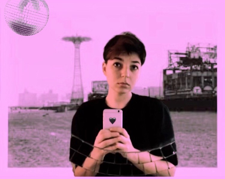 NM Esc is standing in front of a pinkish-purple image of Coney Island with a disco ball in the upper left corner. They are wearing a black t-shirt, and holding an iPhone with a black heart sticker on it that they are using to take the picture.