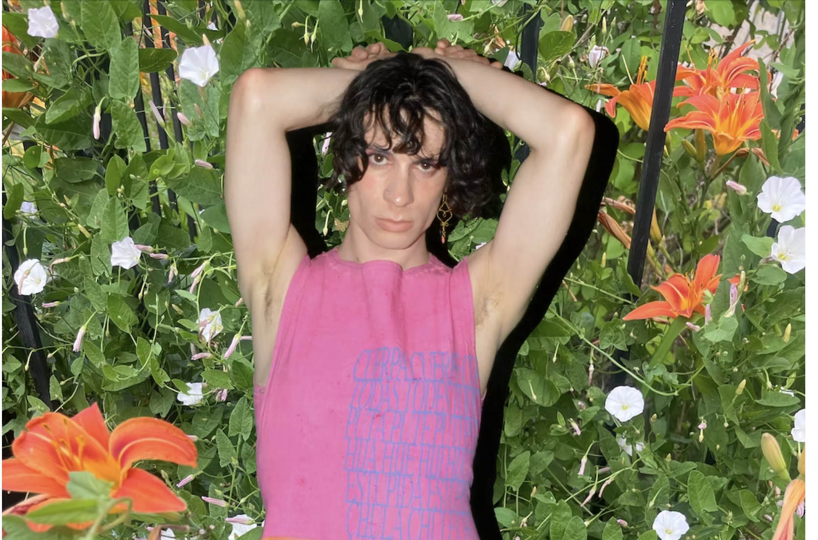 Noa Micaela Fields, a white trans poet, poses with her hands behind her head in front of a verdant floral backdrop. She is wearing a pink cropped top and blue pants silk screened by Mariposa Divina.