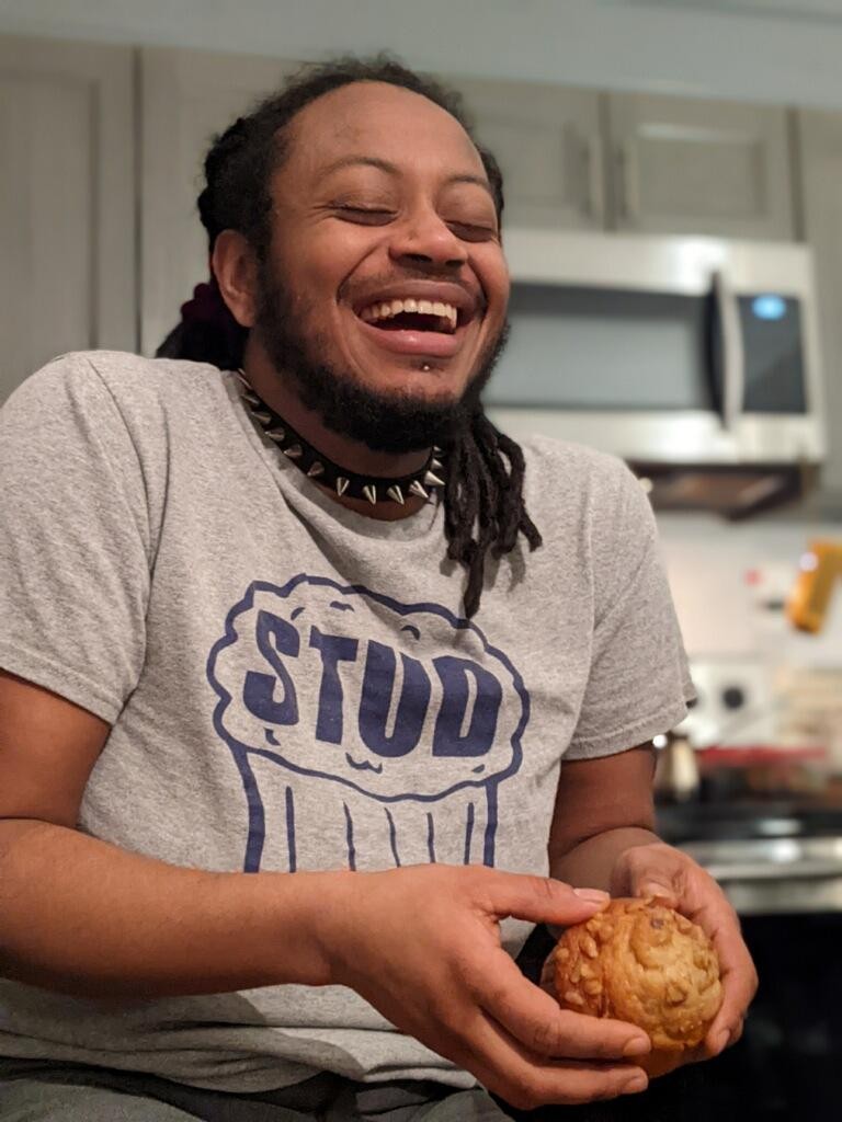 Noor sits in a kitchen, wearing a t-shirt that says stud muffin, laughing