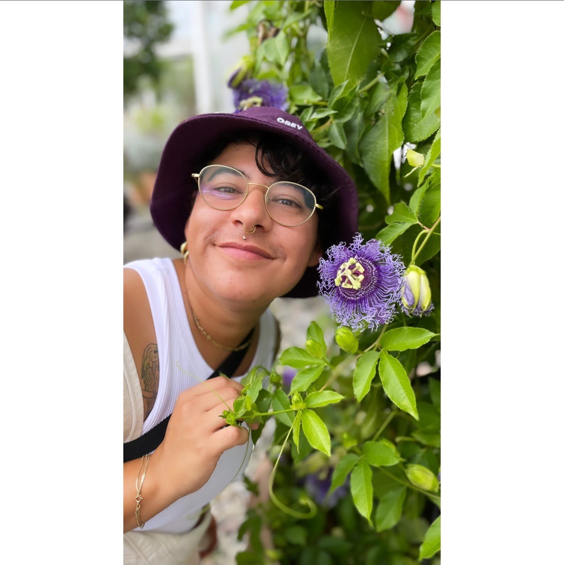 Pao Lebrón smiles next to a purple flower growing on a vine, wearing a purple bucket hat