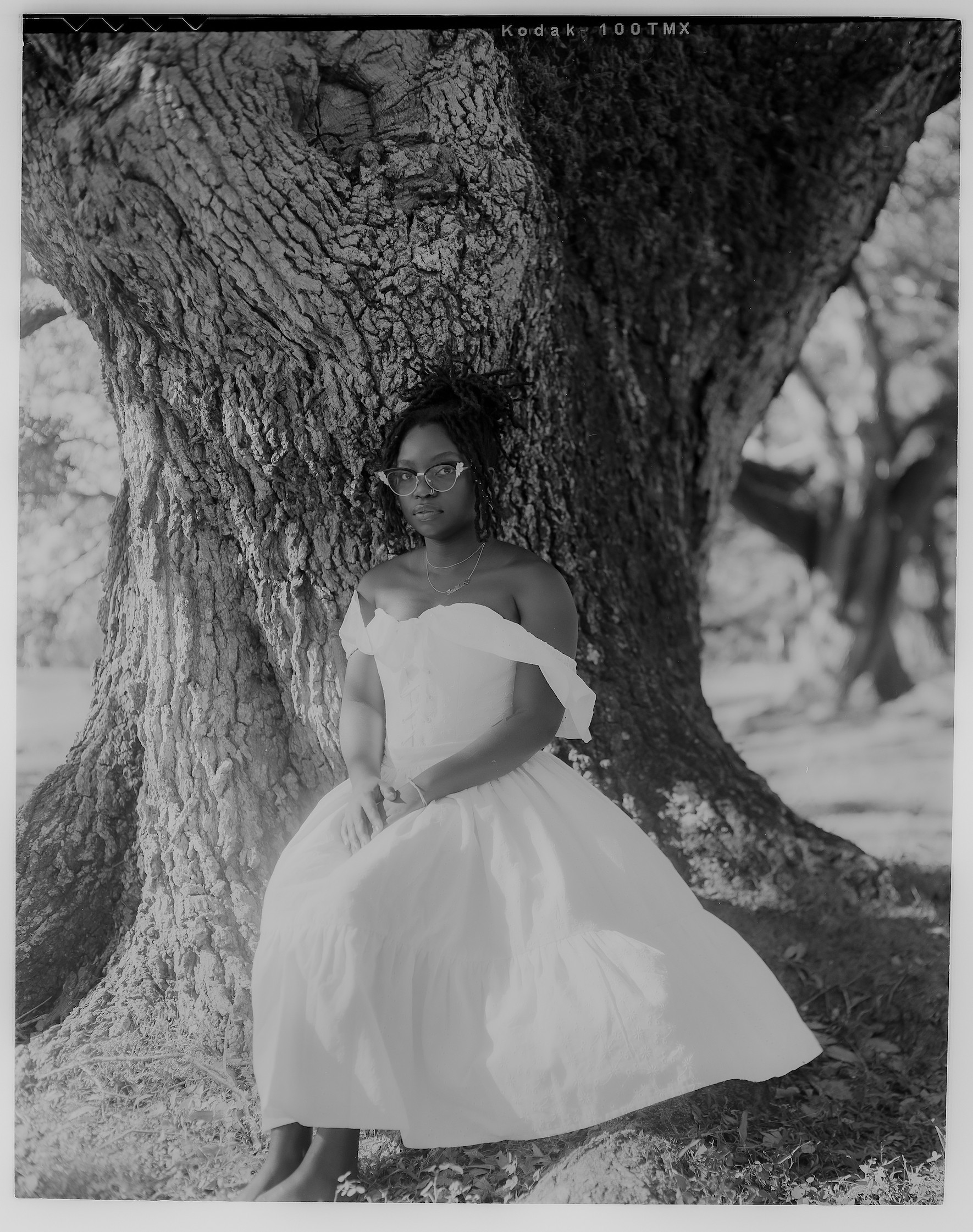 In this black and white portrait, Ra Malika Imhotep sits at the base of an enormous tree, wearing a long white off-the-shoulder dress and cat eye glasses