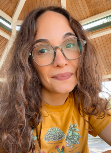 Rachel is featured in a tightly-cropped portrait-oriented photo, seated at what appears to be a picnic bench underneath an umbrella. The photo is cropped from the waist up and to her shoulders and to just above her head. She is wearing an orange t-shirt with illustrations of different cacti and succulents, large square-round green enamel glasses, and her wavy brown hair is loose past her shoulders. She is looking at the camera and smiling.