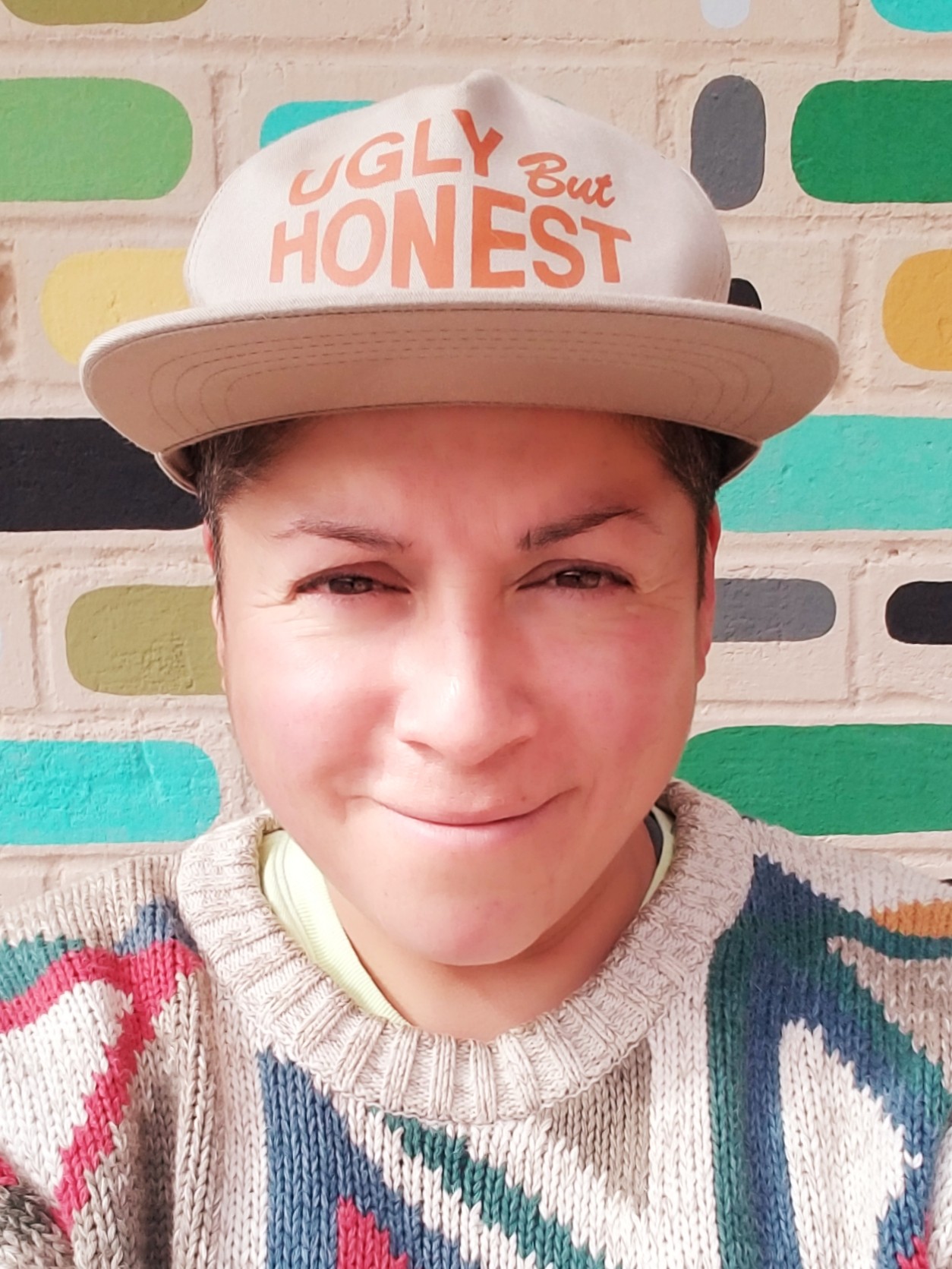 Raquel Gutiérrez is smiling, facing the camera, wearing a baseball cap that reads "Ugly But Honest."