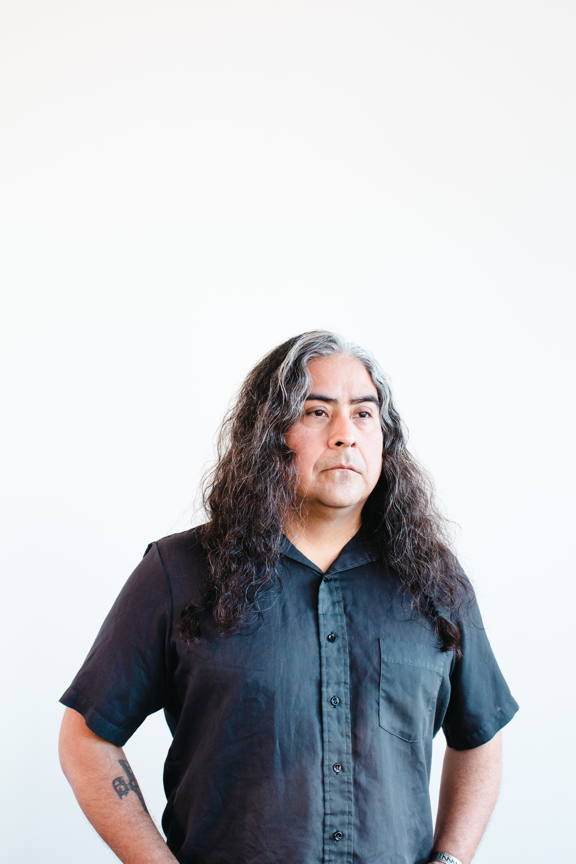 Raven Chacon stands centered in the photograph, cropped from the stomach up. His body angled slightly to the right, the direction in which his eyes are also looking. The backdrop is entirely bright white. Raven wears a short-sleeved button down, his long, wavy, salt-and-pepper hair just beyond his shoulders.