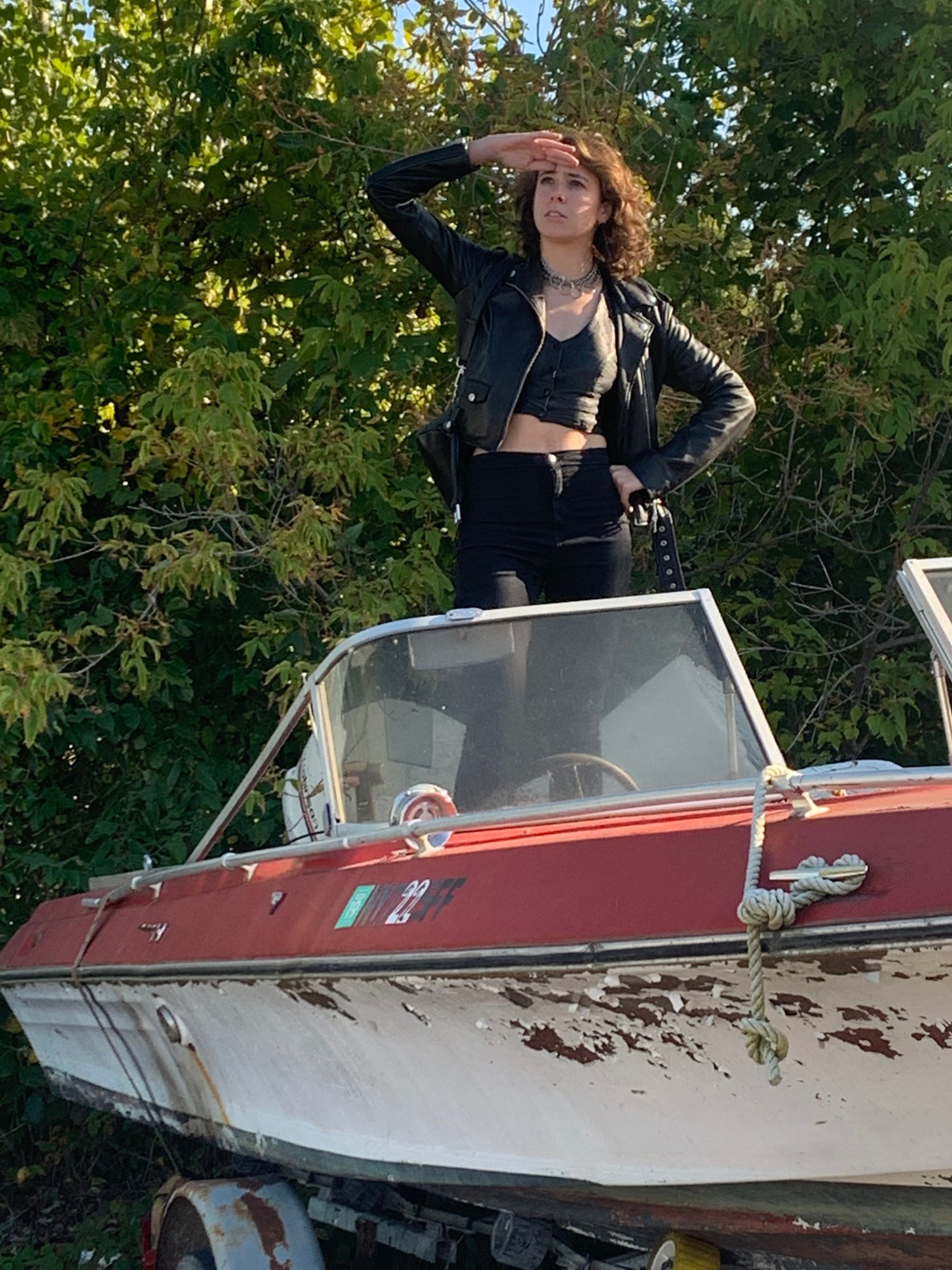 Rebecca Teich is dressed in all black, standing on a dilapidated boat parked on land, with green trees in the background, their hand raised to their eyebrows as if navigating.