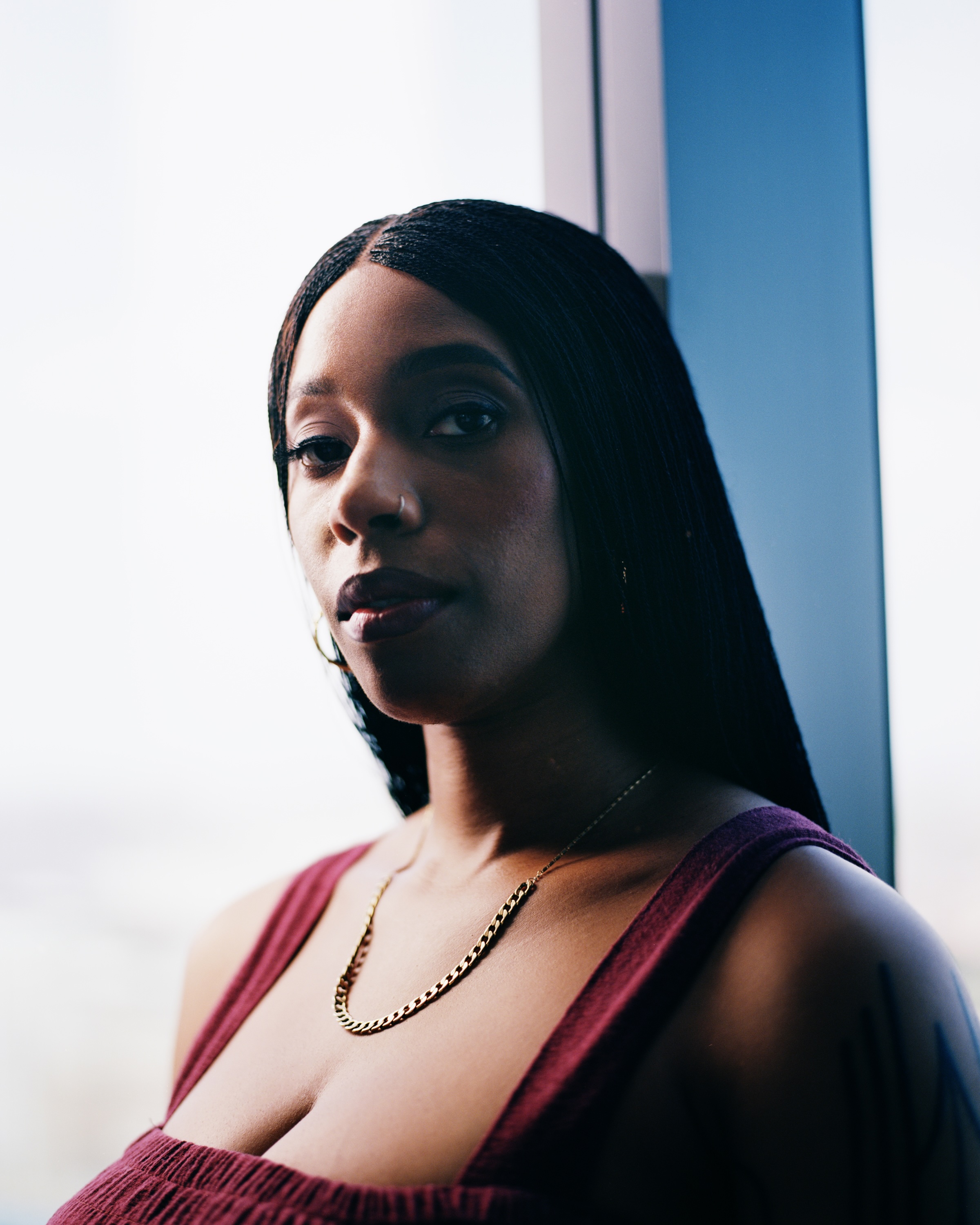 Rena, a lighter-skinned, Black woman with long, tiny twists in her hair, is pictured in front of a window with a partial view of a window pane on a bright day. She wears thick strapped tank top in a maroon color. She is wearing a thin gold chain around her neck and gold hoop earrings. On her arm closest to the camera, is a silhouetted view of a tattoo.