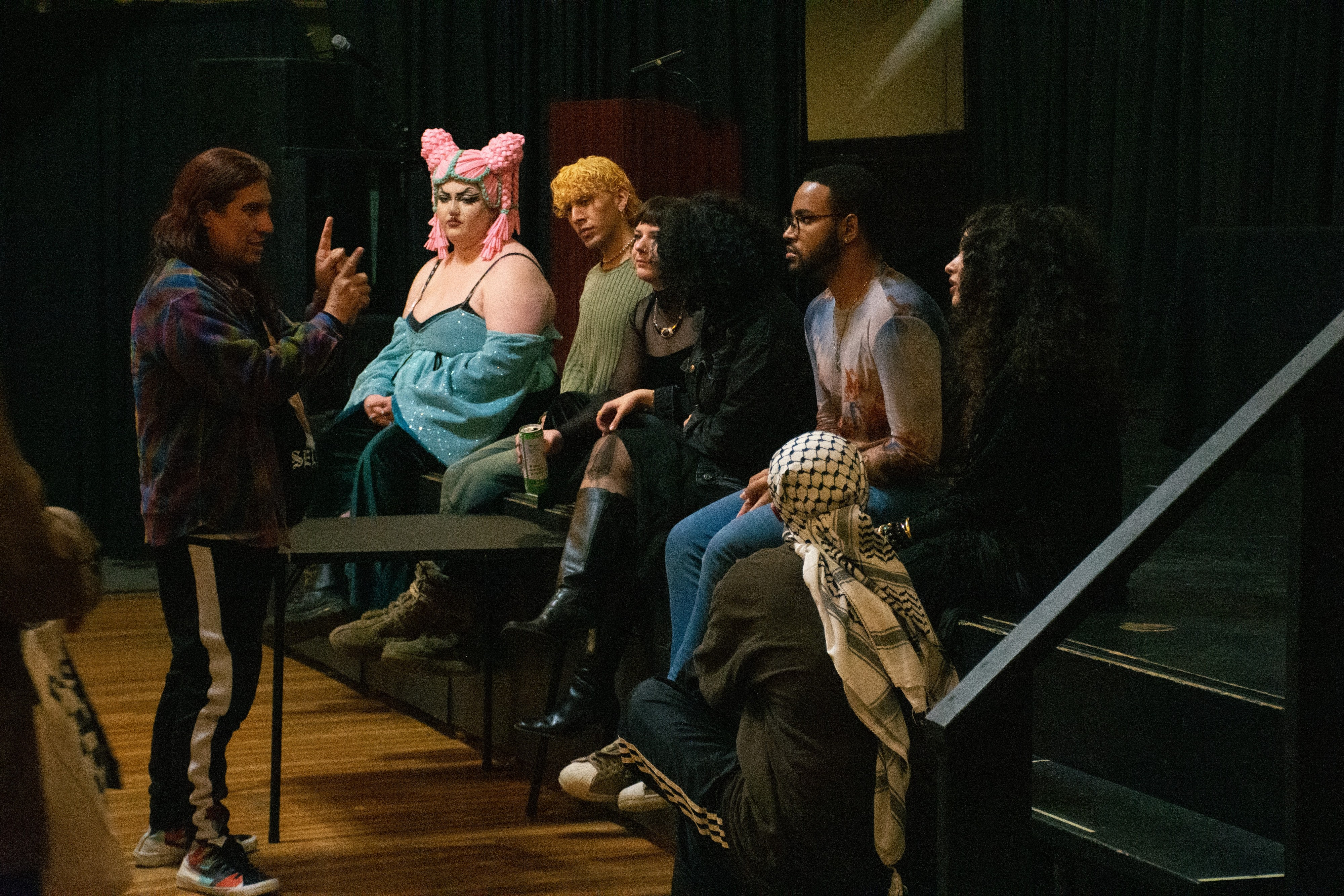 A person gestures with their hands before five individuals sitting on the edge of a stage, while another person wearing a kufiyah scarf on their head kneels to the side.