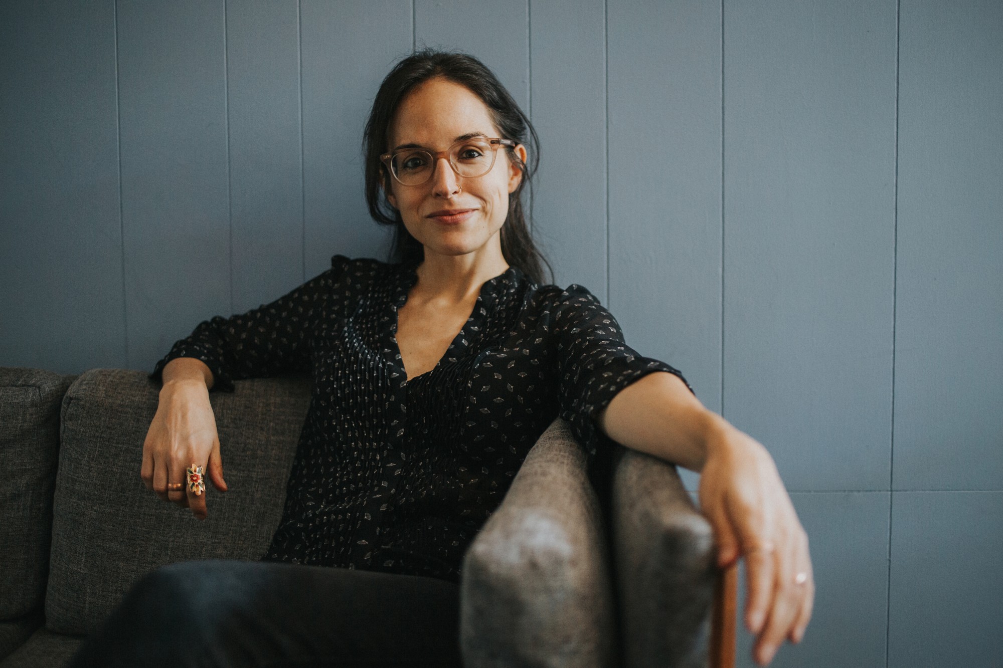 Robin, a white woman with long brown hair, is sitting on a gray couch against a blue background. She wears clear-framed glasses and a black V-necked blouse stamped with a white pattern. One arm rests on the back of the couch, while the other is extended along the armrest. She wears copper rings on several fingers.