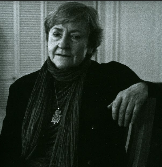 In this black and white portrait, Rosmarie Waldrop sits in a chair with her arm resting on its back, looking into the camera
