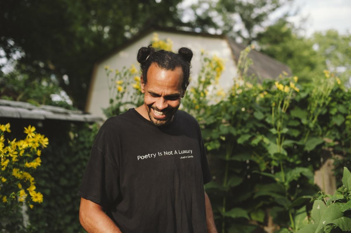 Ross Gay is standing in front of tall plants with yellow flowers, looking down and smiling, wearing a t-shirt that reads "Poetry Is Not A Luxury - Audre Lorde"