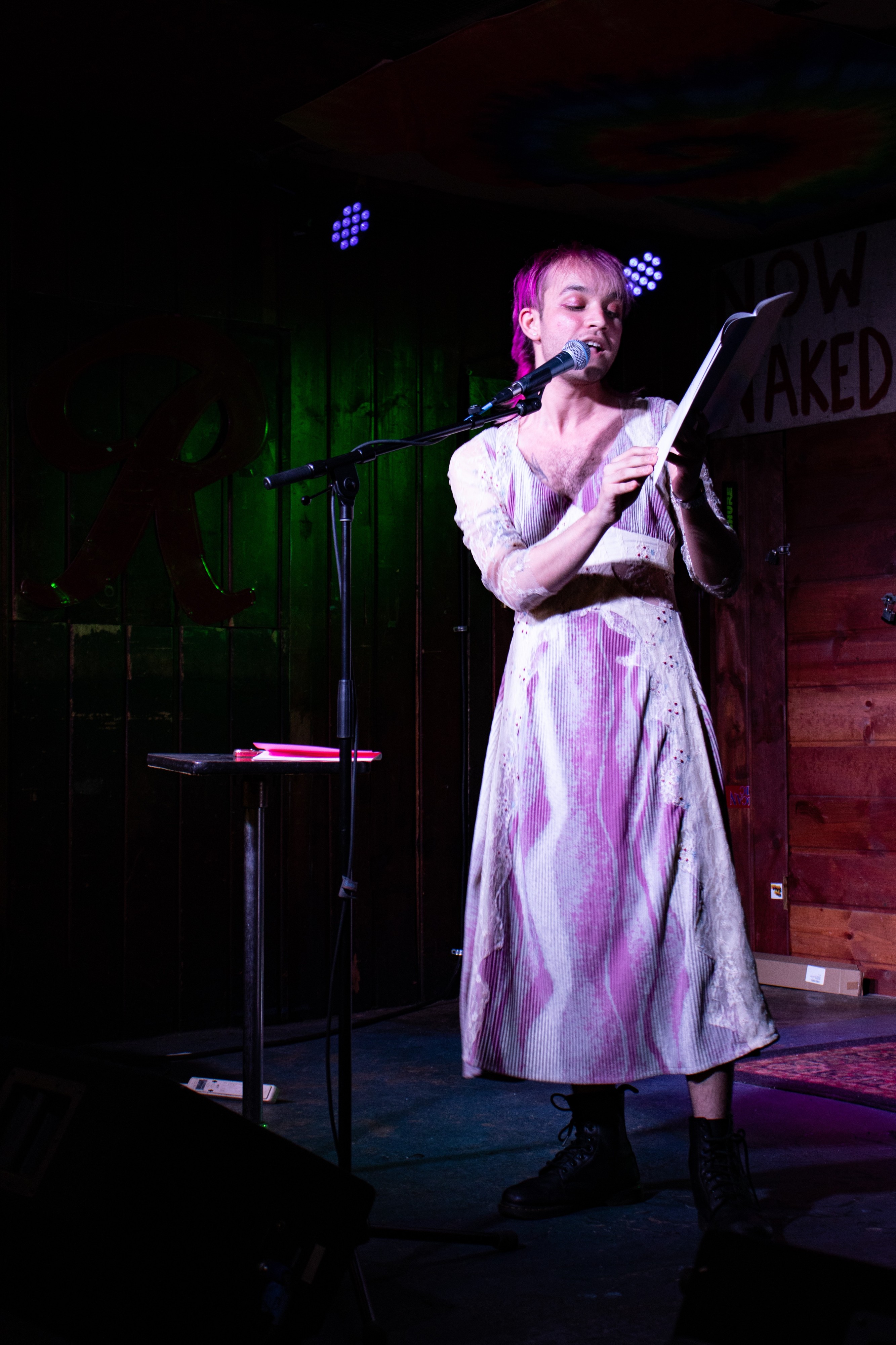 Image description of a Stage at a local Bar in Seattle called the Blue Moon. The pink haired speaker, Ryan Cook, is reading from a book into a large microphone and looking off to the right. they are wearing a wavy pink and white dress with lacy sleeves, as well as black Doc Martens.