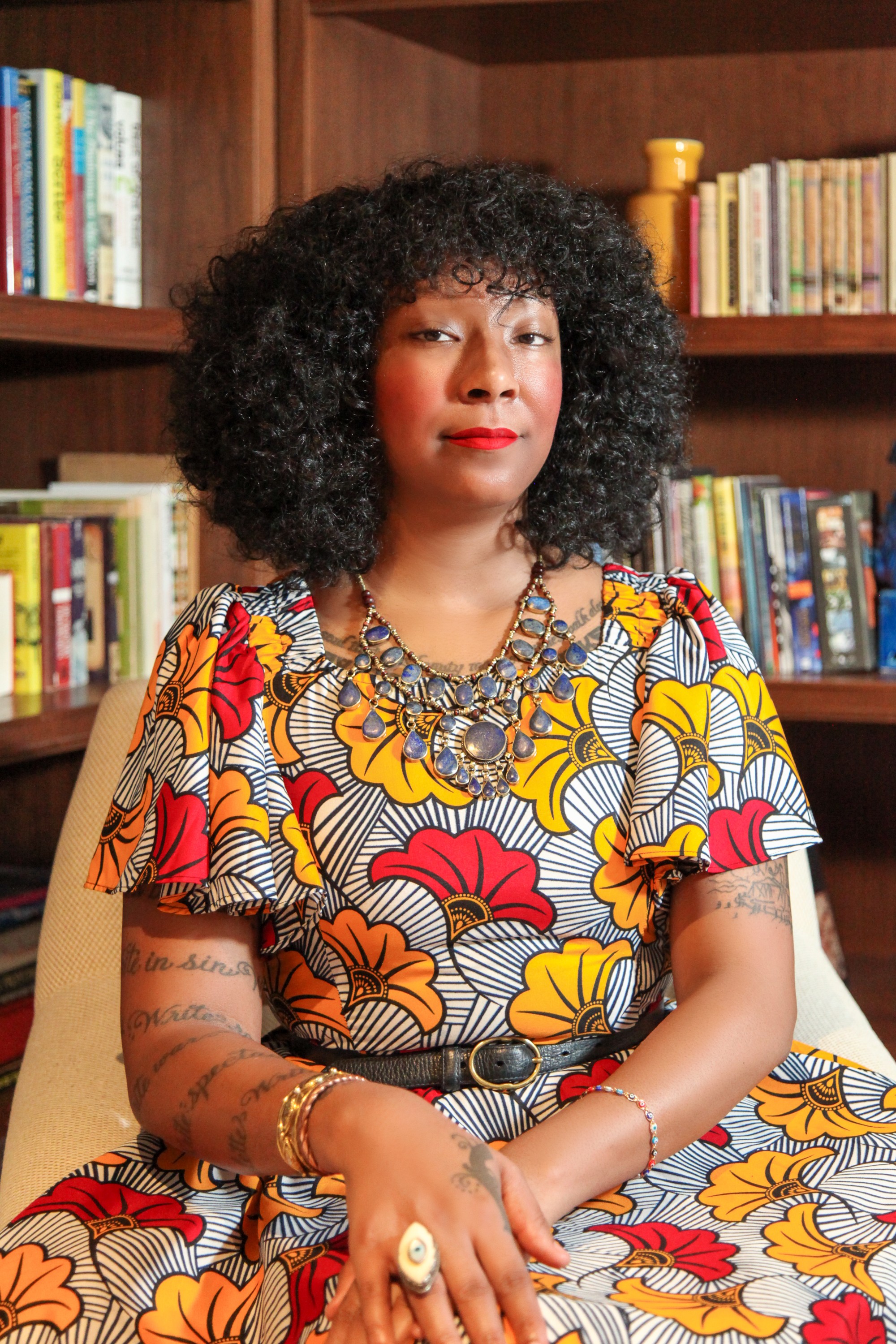 S. Erin is a Black woman with lush curly black hair and red lipstick, seated and framed by shelves of books. She is wearing an Ankara waxcloth navy blue & white striped dress with ruffles and bright red and orange printed fans, along with gold, silver, and lapis lazuli vintage jewelry.
