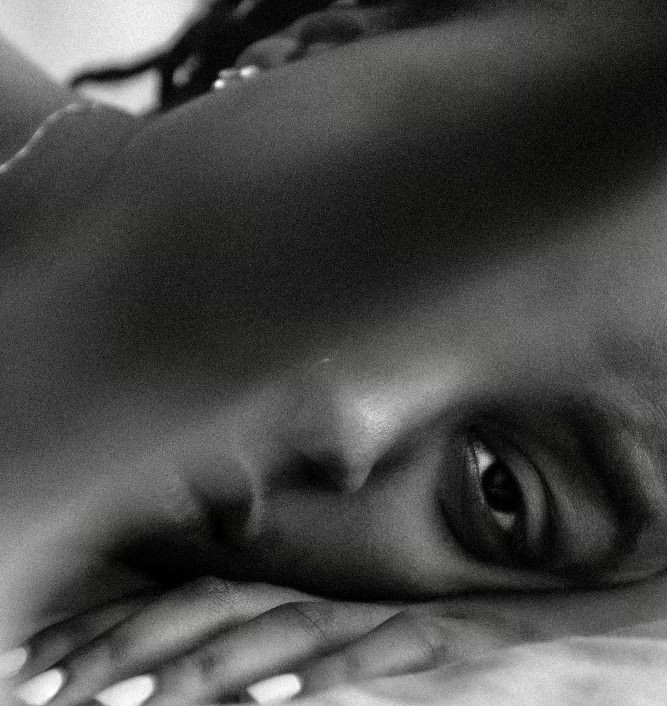 sadé's finger obscures a black and white image of sadé lying down with her face against her hand. she is looking directly into the camera.