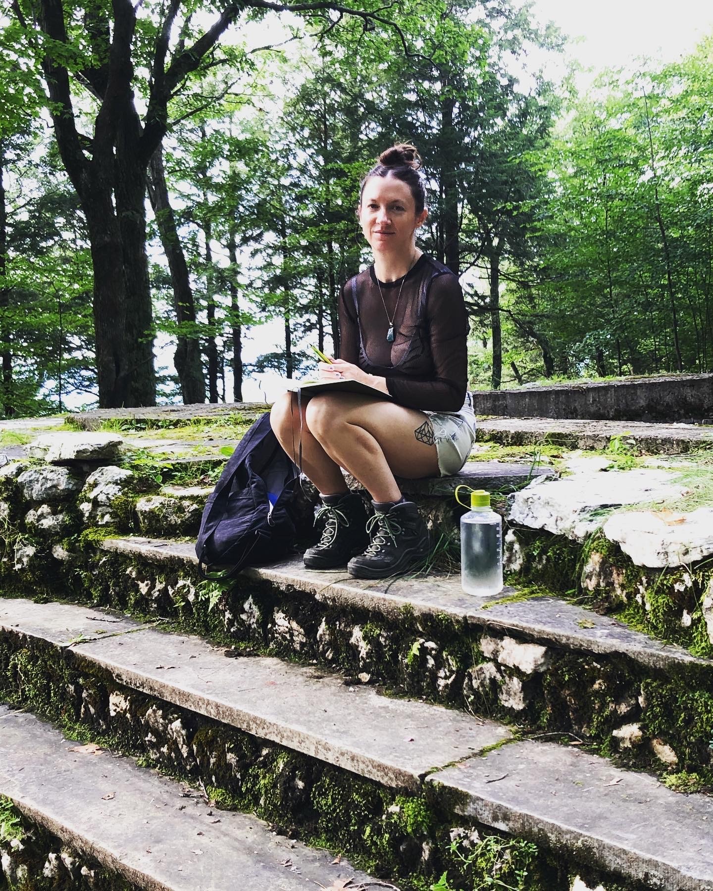 Sara Jane Stoner is featured centered in a portrait-formatted photograph, seated on stone stairs in a park. Her knees are up and placed on them are a book. To her left is a water bottle, to her right a backpack. She is wearing a denim skirt, a black shirt, and smiling at the camera. Behind are trees.