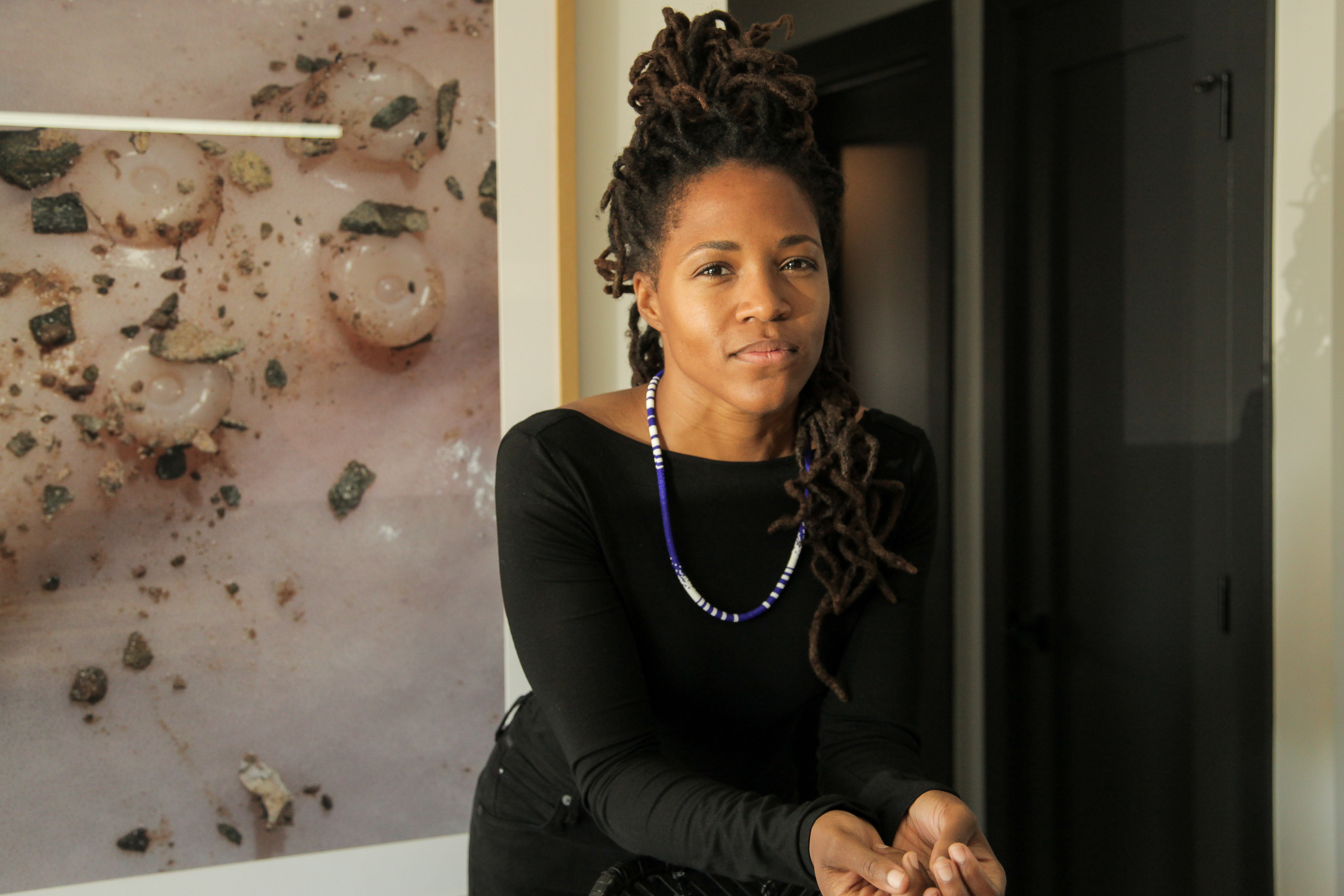 Saretta, a young black woman in a black shirt and Blue and White beaded necklace, leans forward resting her elbows on the back of a stool. She almost smiles. Behind her hangs a large photograph of a squid tentacle.