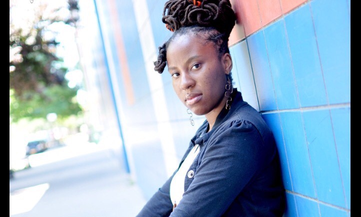 Sherese Francis is featured in a landscape-oriented photograph, cropped from the chest up. The photo is closely cropped but it appears that Sherese is crossing their arms in front of their chest. They are leaning on a wall of light blue and light red tiles. The depth of field is focused closely on Sherese so the background is blurry, but it appears they are outside on what looks like a city street. they are wearing a white t-shirt with an indigo button down over it. They are wearing long dangly wire earrings in a wavy design.