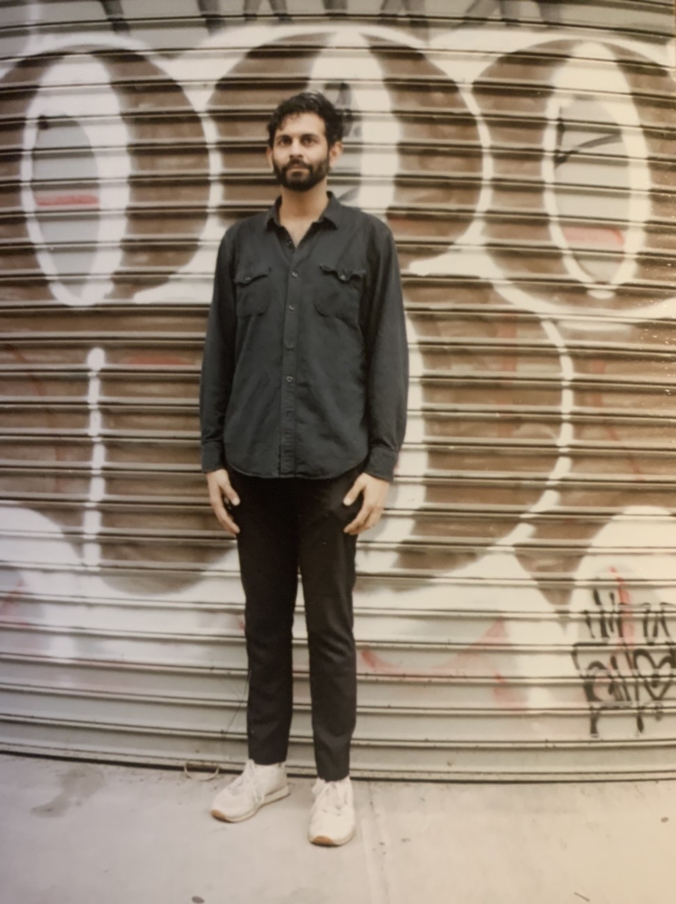 Shiv stands in front of a corrugated metal gate with graffiti on it, wearing all black with white shoes.