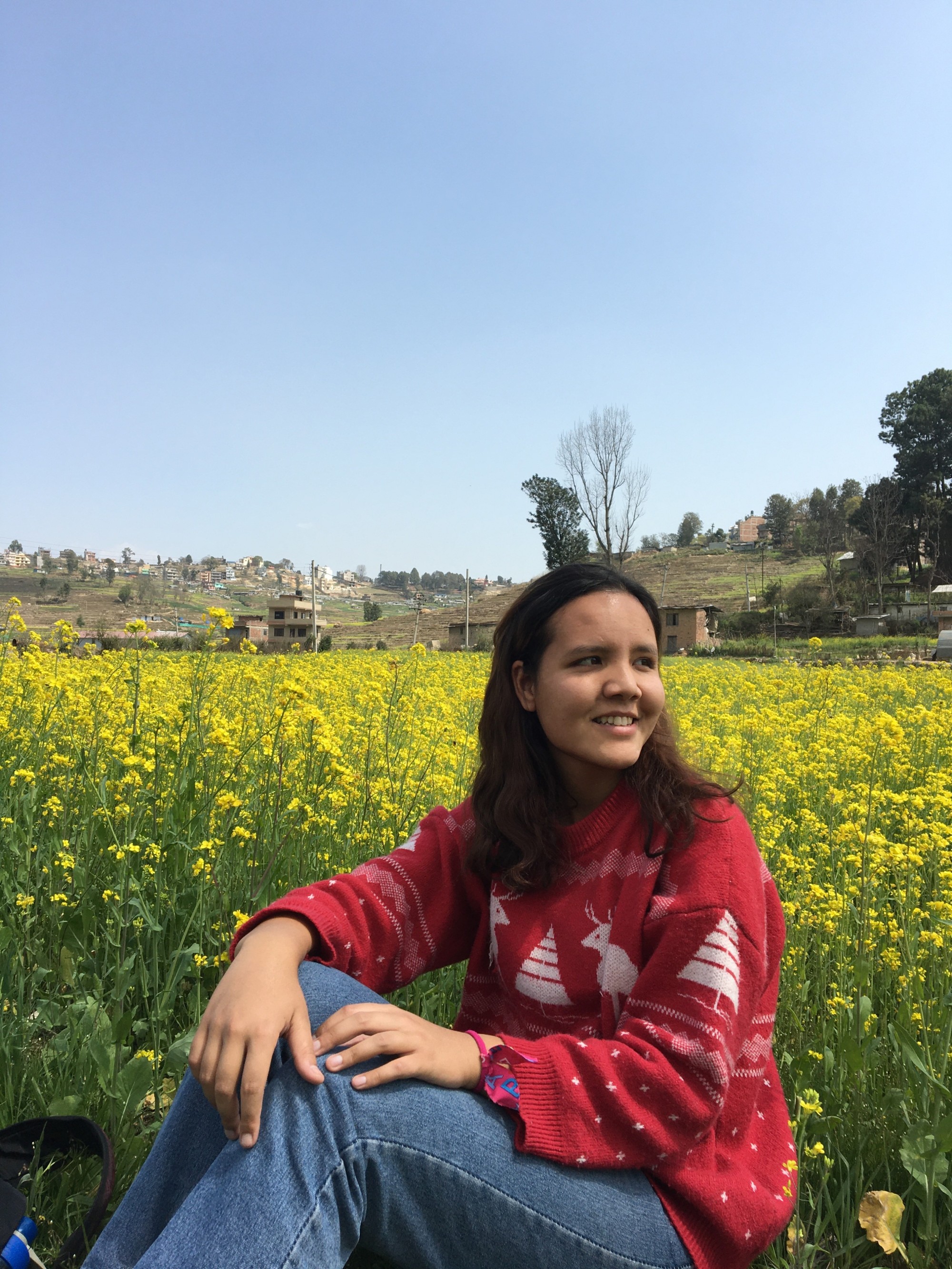 Shuvangi Khadka sits in a field of yellow flowers under a blue sky, wearing a red sweater and smiling, looking away from the camera